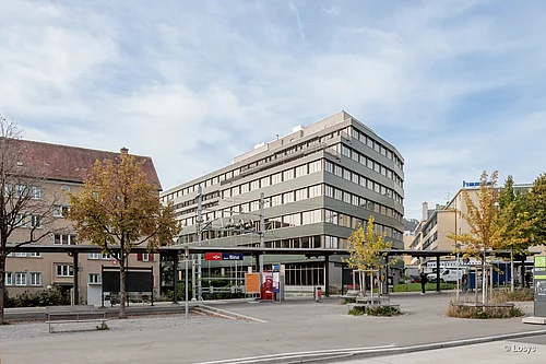 Town square on Binzstrasse, Zurich: in the foreground, a covered ‘Binz’ tram stop with benches and trees in autumnal colours; in the background, a multi-storey grey office block with wide bands of windows and large glass entrance façades, and a paved forecourt.