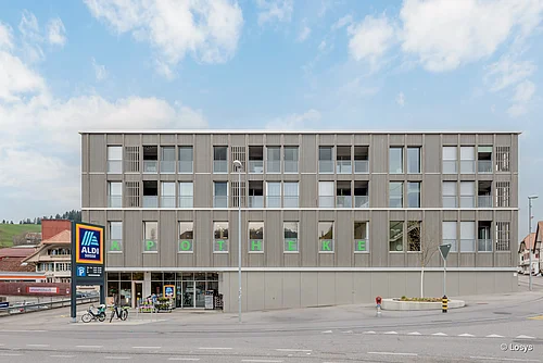 Dreigeschossiges, modernes Wohn‑ und Geschäftshaus mit hellgrauer Holz/Metall‑Fassade. Erdgeschoss: Supermarkt‑Eingang (ALDI) mit automatischer Glasschiebetür und grüne Schrift «APOTHEKE» über Schaufenstern. Obergeschosse: gleichmäßig angeordnete Fenster, Balkone und Rollläden. Straße und Fahrradparkplätze im Vordergrund.