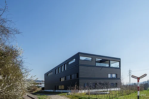 Edificio aziendale rettangolare a tre piani con rivestimento orizzontale grigio scuro, finestre strette e orizzontali e una loggia angolare vetrata; in primo piano un prato, dei cespugli e un binario ferroviario, con un cielo azzurro e limpido.