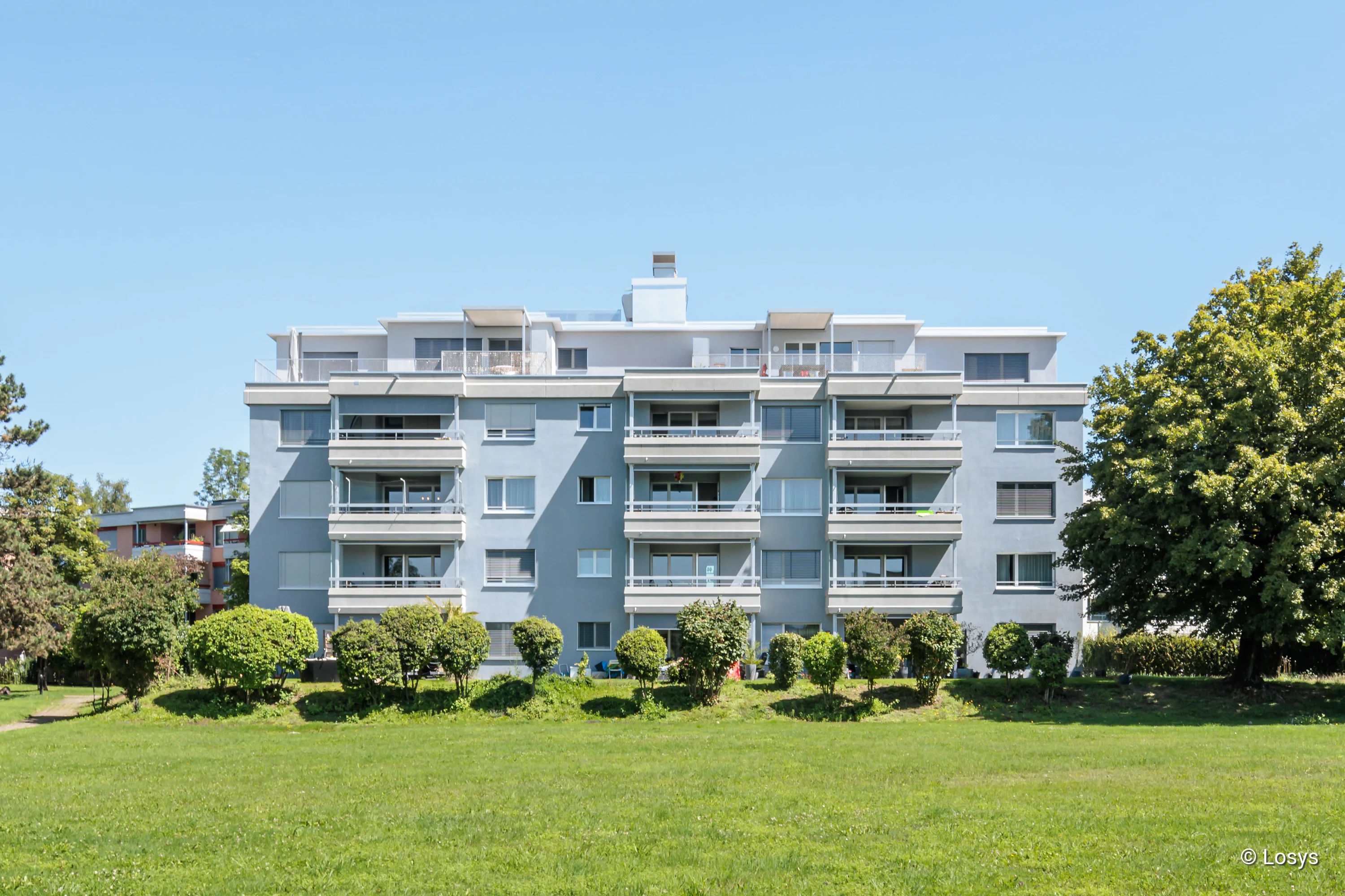 Front view of a light blue block of flats (4–5 storeys) with continuous balconies and glazed balcony doors/sliding glass doors; in the foreground, a large lawn and trees under a clear sky.