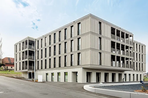Vista esterna del nuovo edificio a quattro piani “Haus T” dell’ospedale di Münsterlingen a Scherzingen: facciata cubica di colore grigio chiaro con finestre strette e a tutta altezza disposte in modo regolare, balconi angolari aperti con parapetti in vetro, piano terra arretrato con diversi ingressi, vialetto d'accesso e piazzale pavimentato in primo piano; sullo sfondo a sinistra, case unifamiliari e cielo nuvoloso. Nessuna persona riconoscibile.