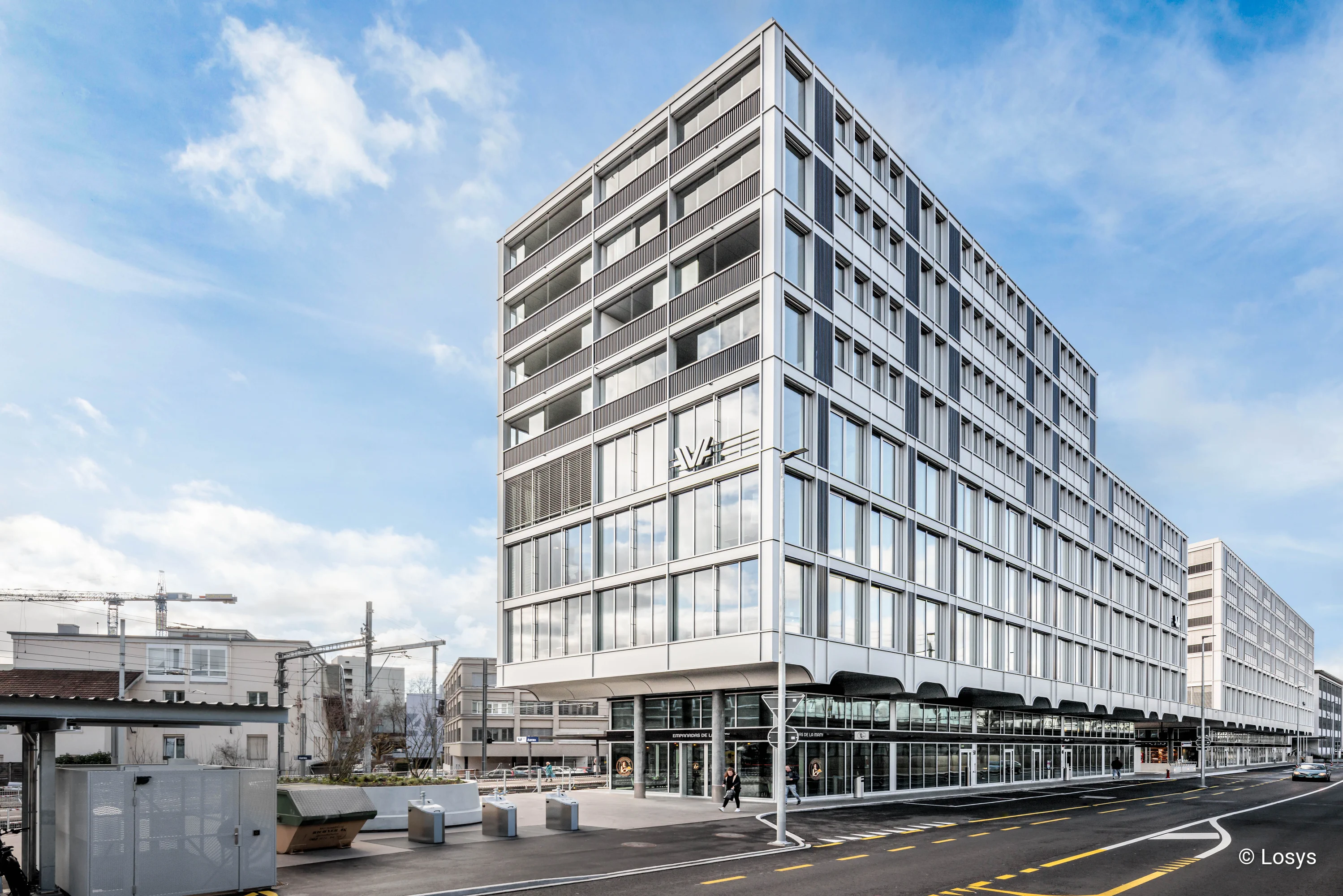 Modernes, mehrgeschossiges Wohn‑ und Geschäftshaus an einer Straßenecke beim Bahnhof Süd Aarau. Helle, rasterartige Fassade aus Metall und großen Glasflächen, bodentiefe Schaufenster und mehrere Eingänge im Erdgeschoss, darüber gleichmäßig angeordnete Fenster und Balkone. Vor dem Gebäude Gehweg, Fahrbahn mit Markierungen und vereinzelte Fußgänger; klarer Himmel im Hintergrund.