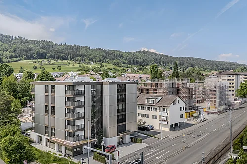 Vue aérienne d'un immeuble d'habitation moderne de cinq étages, avec des panneaux de façade clairs et plusieurs balcons ; au centre, une baie vitrée sombre faisant office d'encorbellement et d'escalier. À droite, un chantier avec des échafaudages ; au premier plan, une rue et une voie ferrée ; à l'arrière-plan, des collines verdoyantes et des maisons individuelles éparpillées.