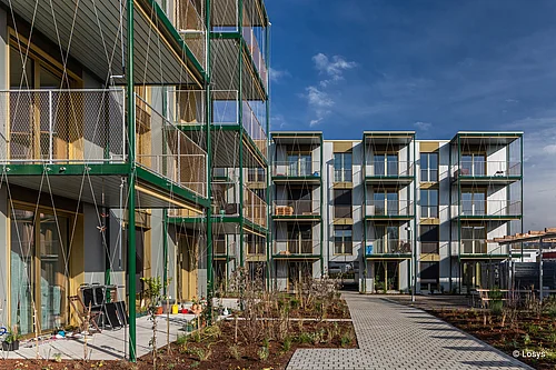 Innenhof eines viergeschossigen, modernen Wohngebäudes mit gestapelten Balkonboxen in grünem Stahlrahmen und Drahtnetz; große verglaste Terrassentüren/ Fensterfronten; Erdgeschoss-Terrassen mit Gartenmöbeln; gepflasterter Weg und neu angelegte Bepflanzung unter blauem Himmel.