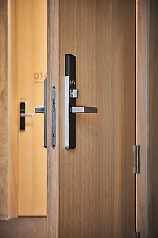 Primo piano di una porta interna in legno, leggermente socchiusa, con finitura effetto rovere, dotata di una moderna serratura elettronica nera e di una maniglia in acciaio inossidabile opaco; sullo sfondo, una porta d’albergo sfocata con il numero della camera.