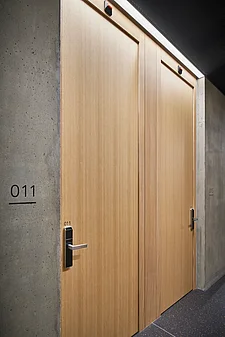 Due porte piatte in legno affiancate nel corridoio di un hotel (camera 011 a sinistra, 015 a destra). Entrambe le porte sono dotate di serratura elettronica a scheda e maniglia a leva in metallo, affiancate da pareti in cemento grezzo, un soffitto scuro perforato e un'illuminazione a filo soffitto continua.