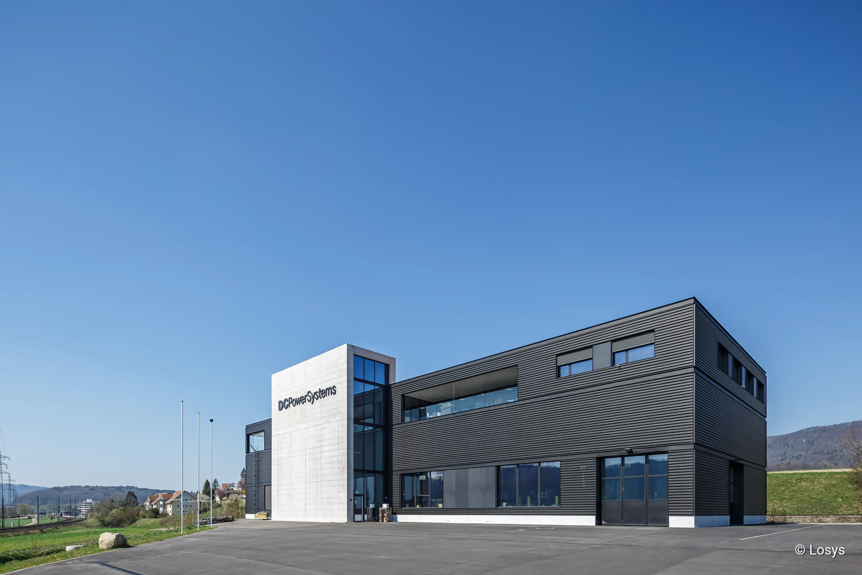 Sede centrale di DCPowerSystems a Otelfingen: torre in cemento bianco, pannelli di facciata scuri, ampie finestre e ingresso vetrato, piazzale asfaltato.