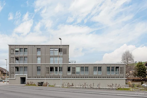 Außenansicht eines modernen, dreigeschossigen Neubaus am Straßenrand mit hellgrauer Fassadenverkleidung aus vertikalen Lamellen, gleichmäßig angeordneten Fenstern mit Rollläden, seitlichem Eingang, kleiner Randbegrünung und Straßenlaternen; klarer bis leicht bewölkter Himmel, keine Personen sichtbar.