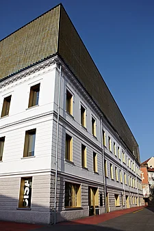 Coin de façade du Palacinema à Locarno, photographié en oblique depuis le bas : Bâtiment en crépi blanc de plusieurs étages, avec des fenêtres disposées de manière régulière et soulignées de jaune ; au rez-de-chaussée, porte(s) d'entrée vitrée(s) et vitrines visibles ; au-dessus, un surmontant cubique en forme de grille, d'un doré éclatant ; ciel bleu limpide.