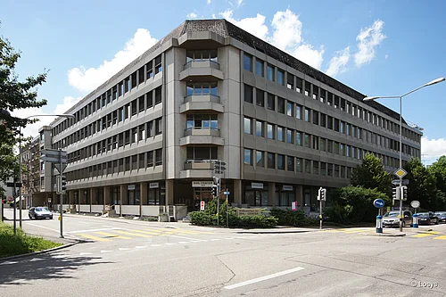 Bâtiment administratif à plusieurs étages situé à un carrefour en angle : façade en béton clair avec des rangées de fenêtres, angle arrondi avec des balcons, entrée de plain-pied avec une enseigne « SIHL-CENTER » et un passage piéton.