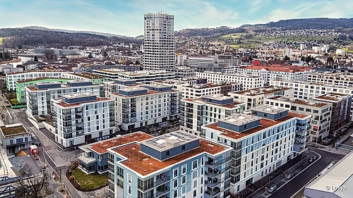 Arbonia-Referenz: Vogelperspektive auf das Wohnquartier Limmatfeld in Dietikon. Mehrere moderne, rechteckige Wohngebäude und ein hoher Wohnturm, helle Fassaden mit großflächigen Fenstern und Glasbalkonen, flache Dächer teils mit Dachbegrünung, erschlossene Straßen, Fußwege und angrenzende hügelige Landschaft sichtbar.