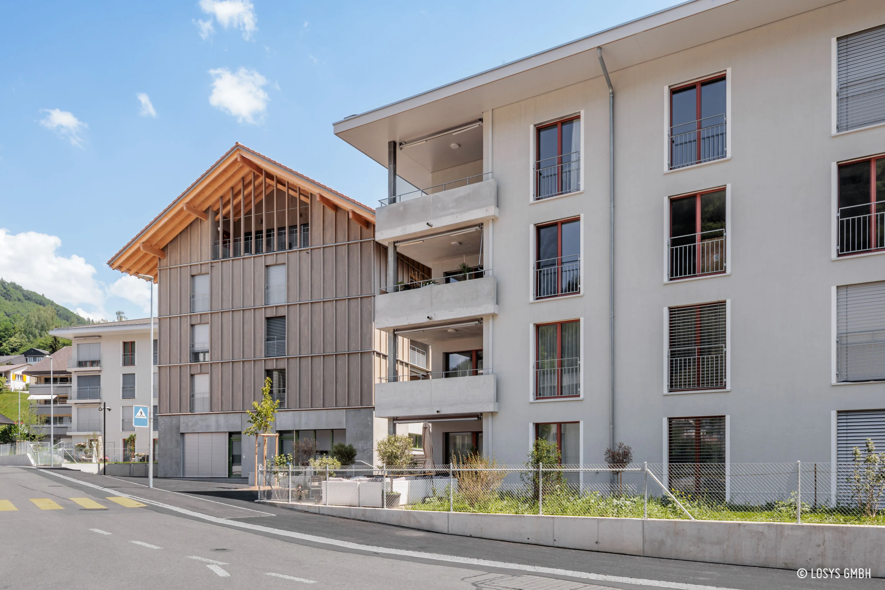 Vue de rue sur deux immeubles d'habitation modernes : à droite, un bâtiment au crépi clair avec des balcons en quinconce, des encadrements de fenêtres rouges et des entrées au rez-de-chaussée ; à gauche, une construction en bois avec un grand pignon, des lamelles de bois verticales et des portes de garage. Au premier plan, un trottoir avec un passage piéton, un panneau « Réservé aux piétons », une clôture basse en grillage métallique et de petites plantations sous un ciel bleu.