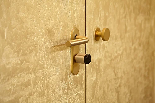 Gros plan sur une porte intérieure à deux vantaux présentant une surface texturée de couleur dorée. Un étroit interstice est visible entre les vantaux ; on distingue clairement une poignée à levier dorée munie d'un bouton cylindrique et un bouton de porte rond ; la quincaillerie et la serrure sont visibles.