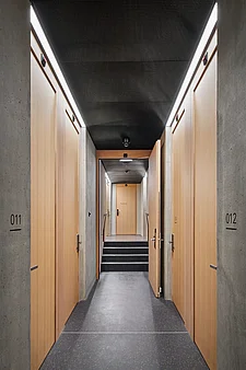 Corridoio dell’hotel con porte delle camere in legno chiaro su entrambi i lati (n. 011/012), quella a destra parzialmente aperta; soffitto acustico scuro con strisce LED, pavimento nero e tre gradini che conducono alla porta “Tecnica”.