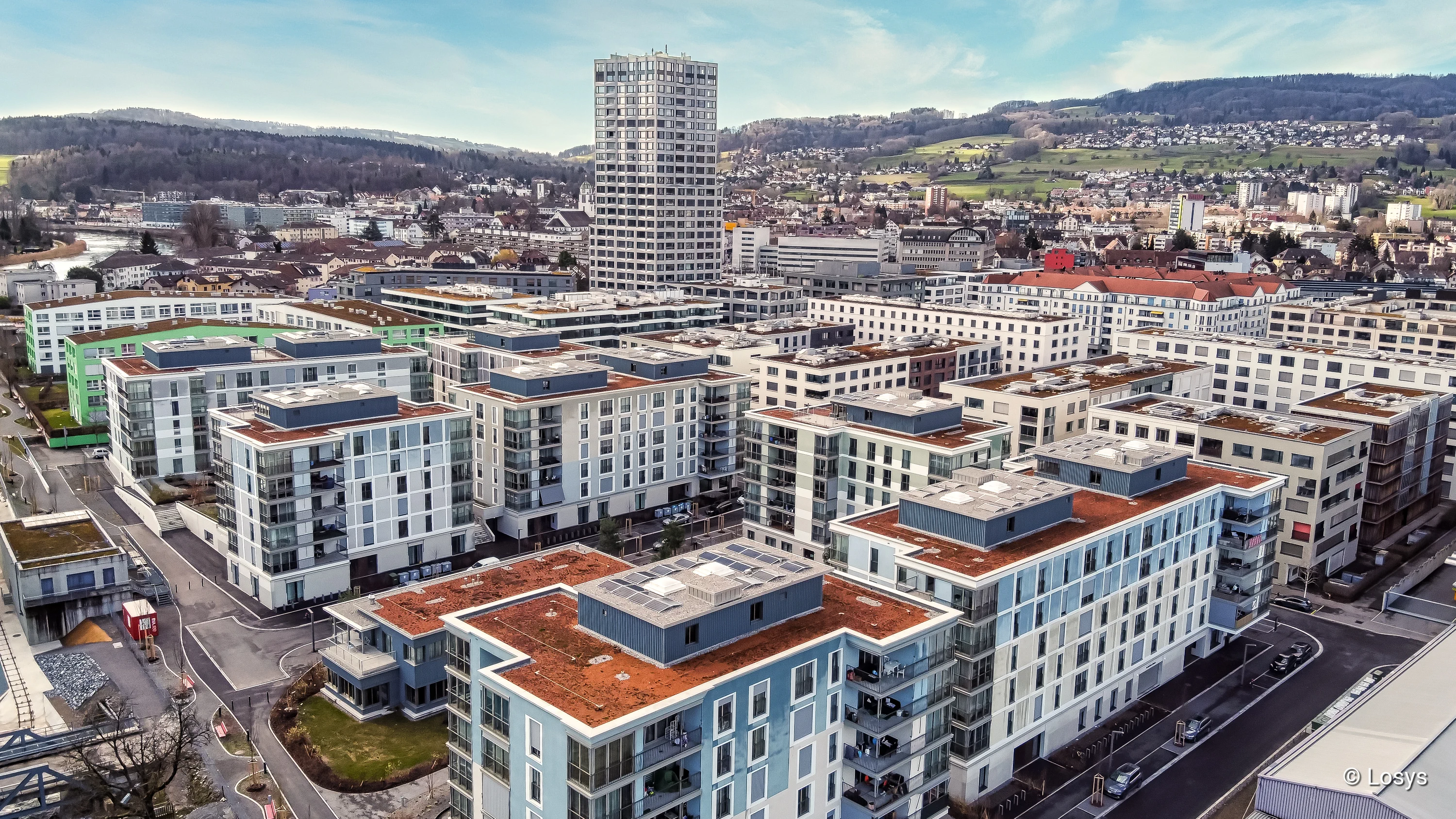 Arbonia-Referenz: Vogelperspektive auf das Wohnquartier Limmatfeld in Dietikon. Mehrere moderne, rechteckige Wohngebäude und ein hoher Wohnturm, helle Fassaden mit großflächigen Fenstern und Glasbalkonen, flache Dächer teils mit Dachbegrünung, erschlossene Straßen, Fußwege und angrenzende hügelige Landschaft sichtbar.