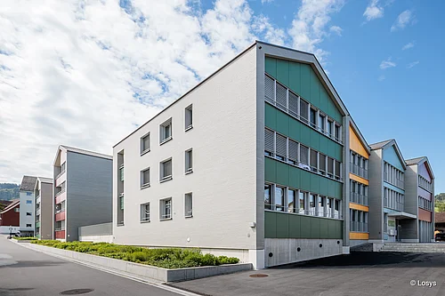 Außenansicht einer modernen Wohnüberbauung in Appenzell: mehrere mehrgeschossige Gebäude mit weißen Backsteinfassaden, farbigen Fassadenfeldern (grün, gelb, rot), zahlreichen Fenstern mit Lamellen, mittigem Vorplatz mit Asphalt und niedrigen Pflanzbeeten. Keine Personen sichtbar.