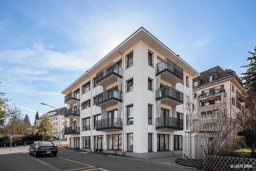 Angolo di un moderno condominio bianco a Zurigo con tre piani residenziali, balconi in metallo scuro su ogni piano, grandi finestre al piano terra, marciapiede e auto parcheggiata.