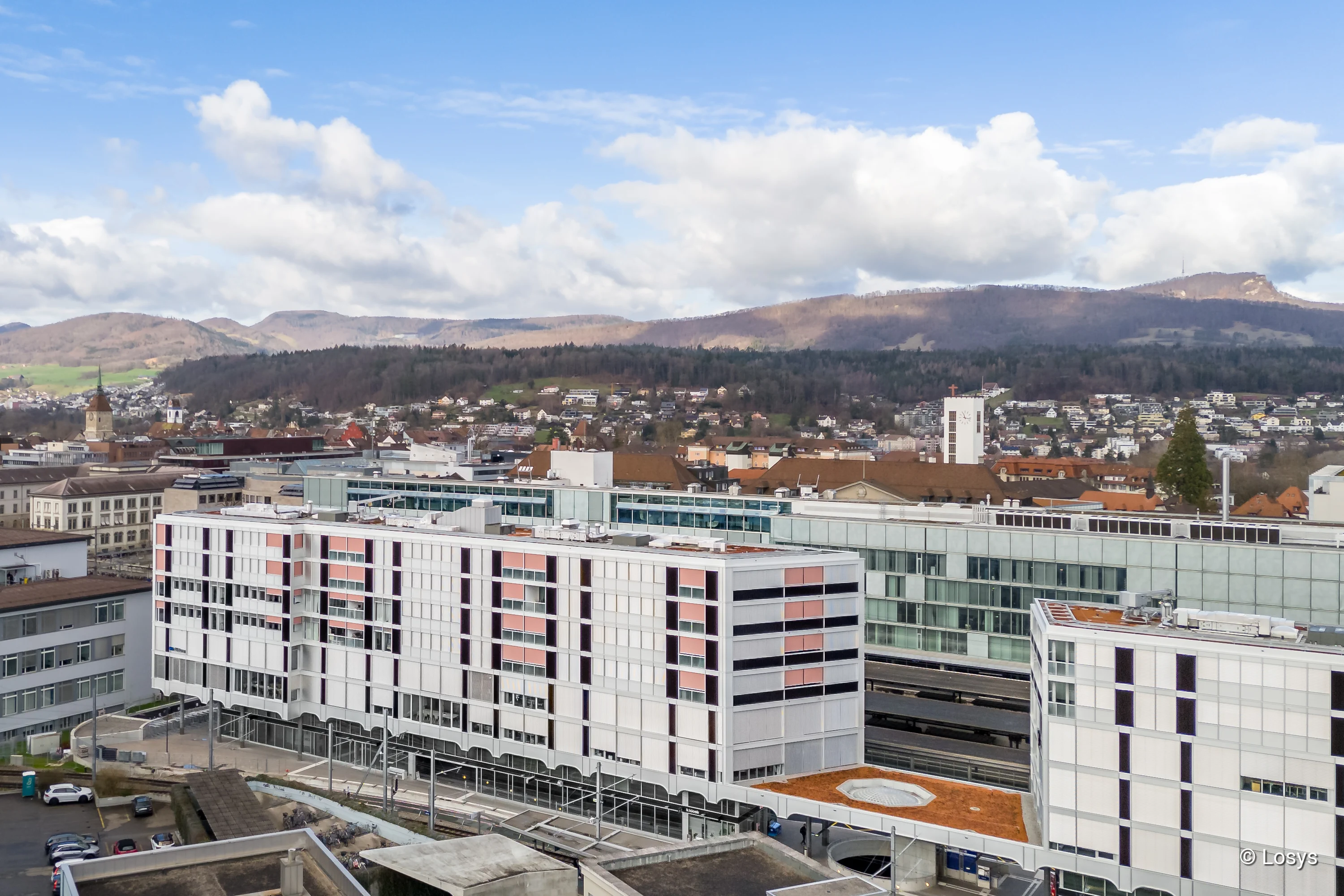 Vogelperspektive auf ein mehrgeschossiges, längliches Wohn‑ und Geschäftshaus am Bahnhof: helle weiße Fassade mit regelmäßigen Fensterfeldern, rosa-farbene Sonnenschutzblenden und dunklen vertikalen Bändern, verglaste Bereiche entlang des Bahnsteigs sowie Erdgeschossöffnungen über den Gleisen. Im Hintergrund Bahnhofsbauten, Stadt und bewaldete Hügel unter blauem Himmel.