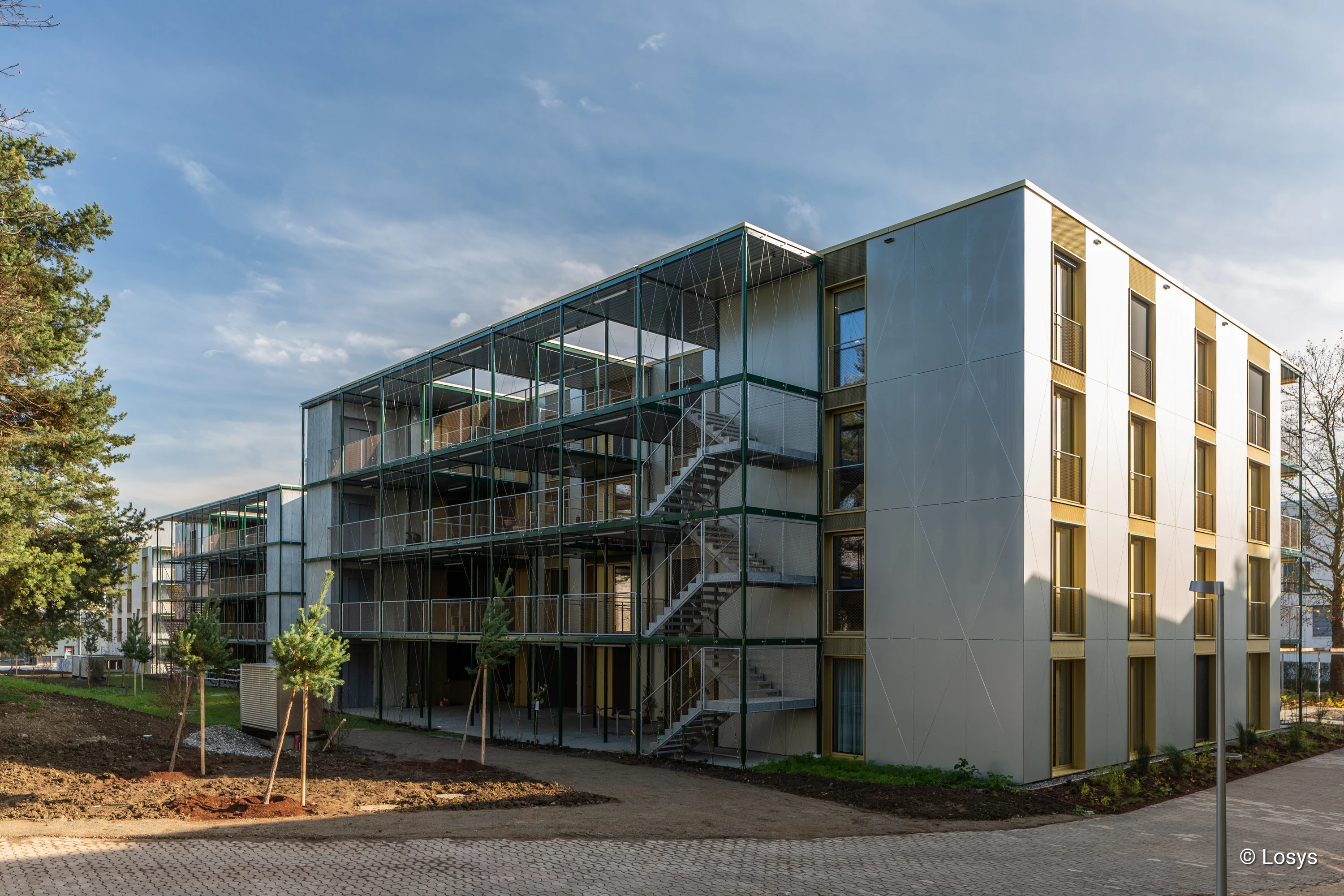 4-geschossiges Gebäude mit Stahlbalkonen und Außentreppen, verglasten Balkonfronten/Glasschiebetüren, Vorplatz und jungen Bäumen.