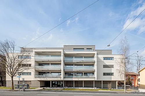 A front view of a modern four-storey residential building: a light-coloured rendered façade, wide balconies with transparent glass balustrades, a recessed ground floor with an open driveway/entrance, large windows and balcony doors, tram overhead lines and bare trees lining the street.