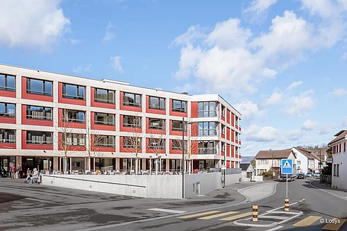 Vista esterna di un moderno edificio residenziale e commerciale di quattro piani (Hauptstrasse 12, Doettingen) con facciata rossa e bianca, ampie vetrate e balconi, terrazza al piano terra, rampa di accesso al parcheggio sotterraneo e attraversamento pedonale; alcune persone in primo piano.