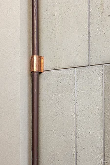 Close-up of a façade detail: on the left, smooth, light beige render on a corner of the building; on the right, light-coloured, slightly textured façade panels with visible joints. A dark, round rainwater downpipe runs vertically and is secured to the cladding with a copper-coloured fixing bracket; this emphasises the details of the materials, colours and fixings.