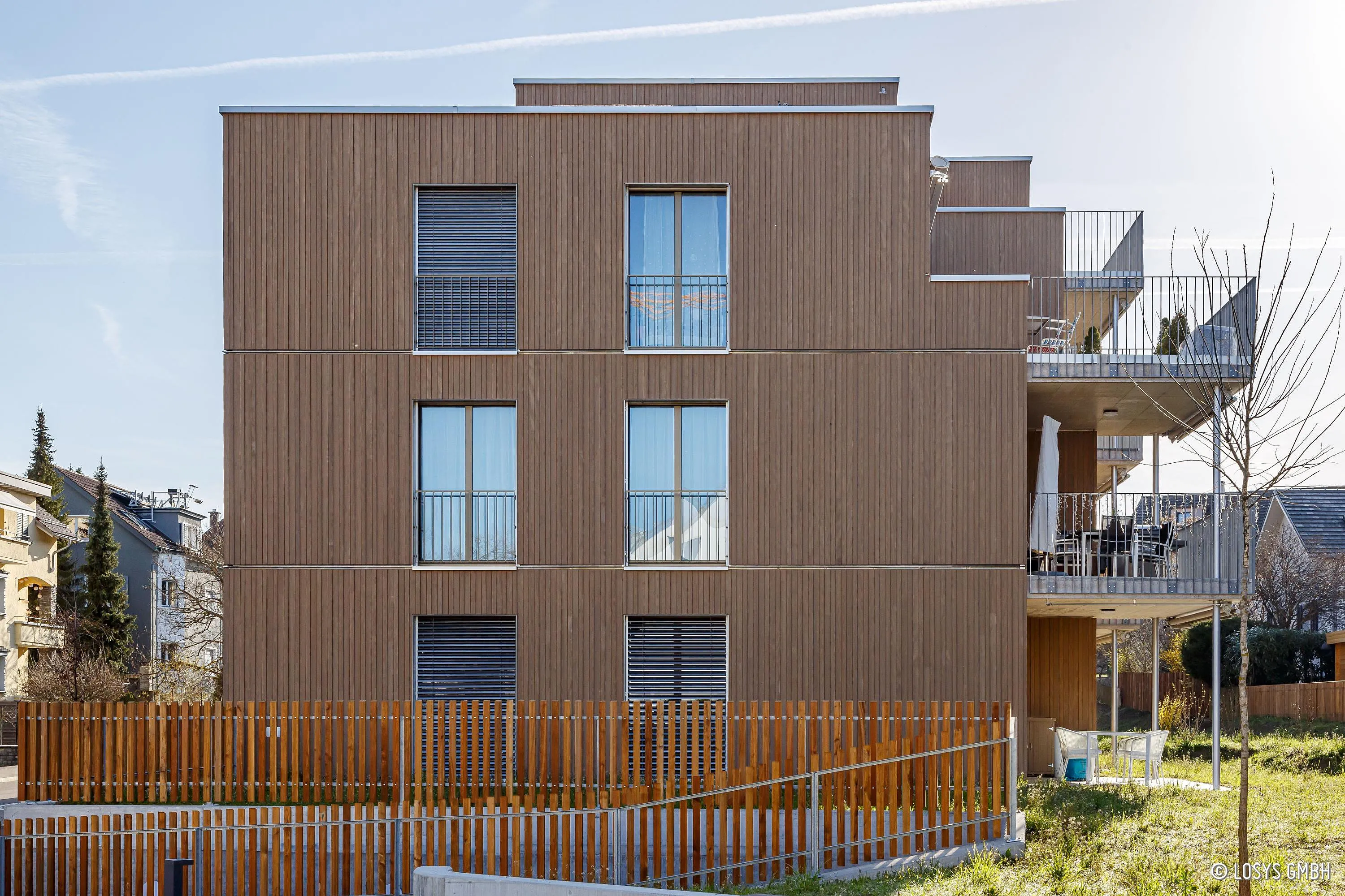 Modernes dreigeschossiges Wohnhaus mit vertikaler Holzfassade, bodentiefen Fenstern und gestaffelten Balkonen rechts; im Vordergrund ein Holzlattenzaun und Rasenfläche.