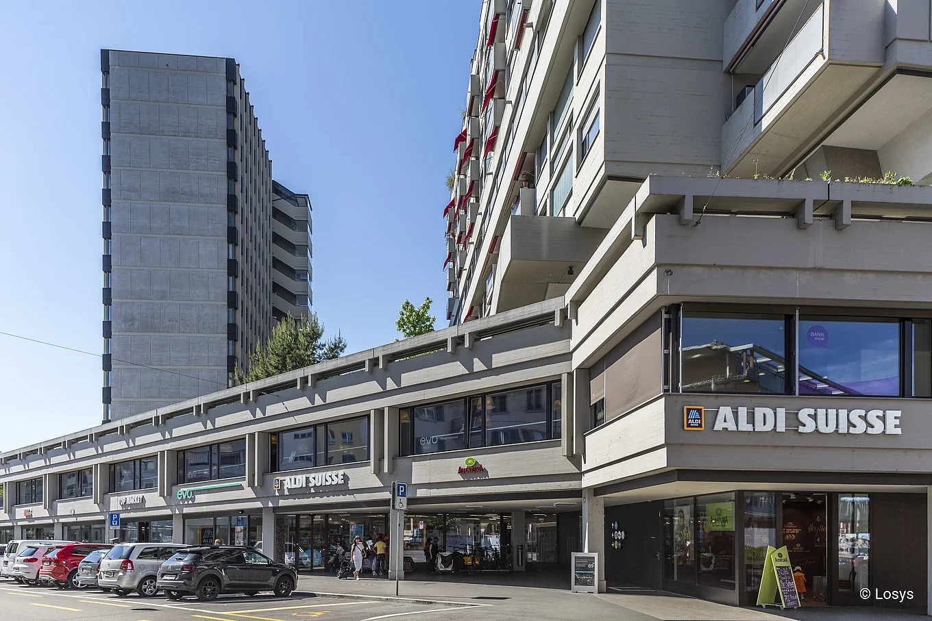 Straßenansicht des Bauhofs Zürich‑Oerlikon: langgestreckter, betongeprägter Gebäuderiegel mit durchgehender Glas-Schaufensterfront und ALDI SUISSE-Eingang im Erdgeschoss; darüber mehrgeschossige Wohnungen mit Balkonen, davor Parkplätze und Fußgänger.