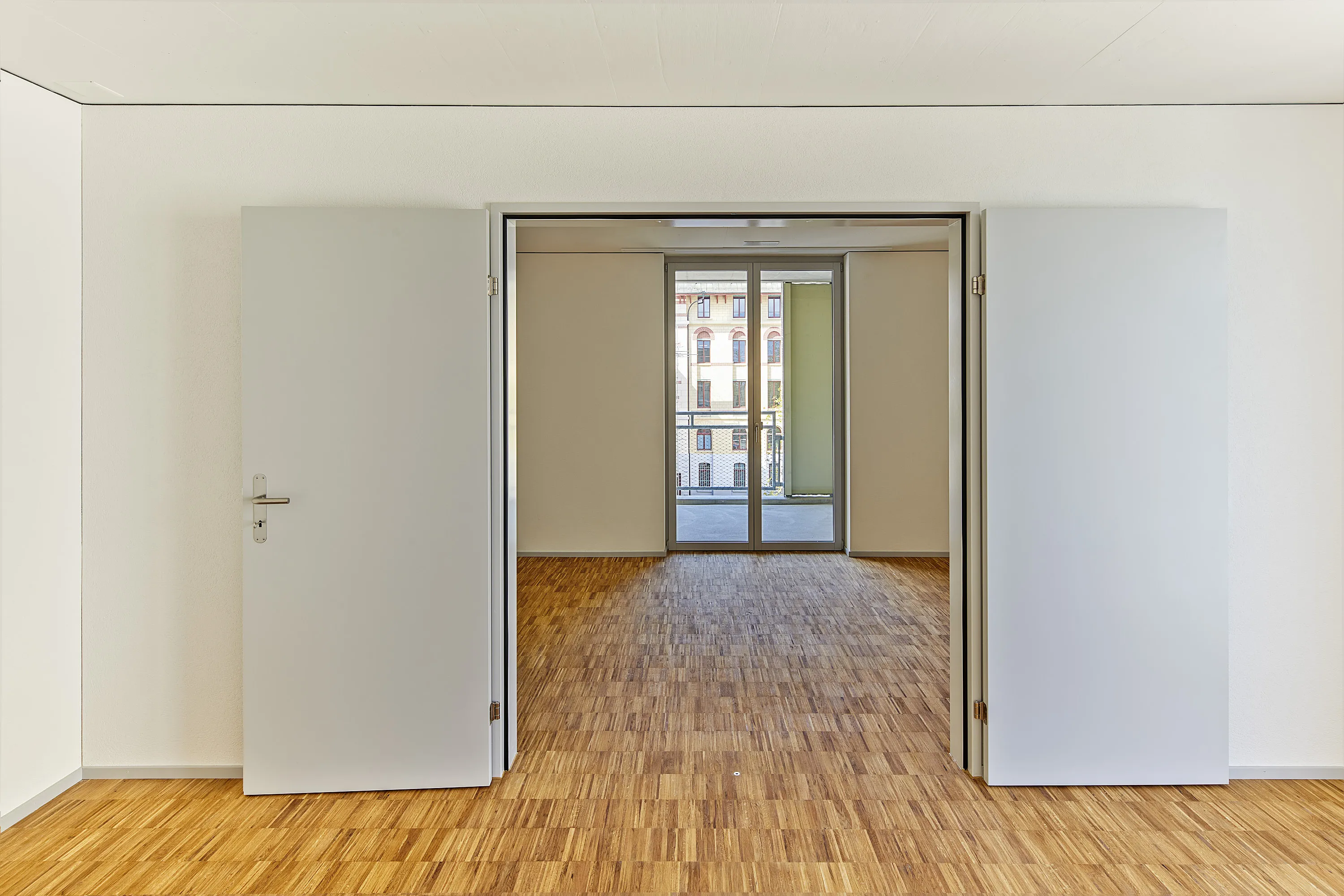 View through open white double doors into two bright, empty rooms with warm parquet flooring; in the rear room a large glass door to the balcony, facades of neighbouring buildings visible outside.