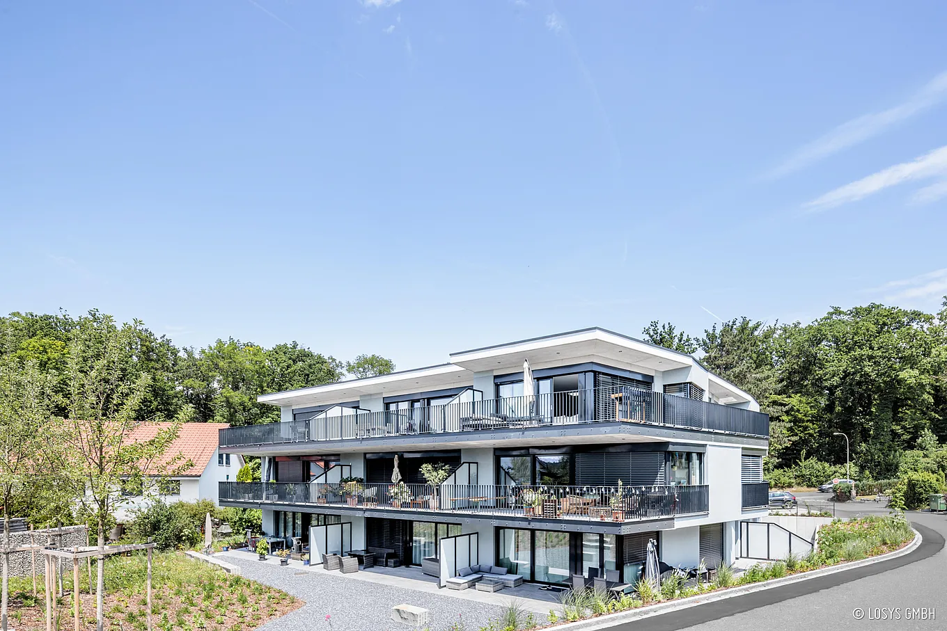 Vorderansicht eines dreigeschossigen, weißen Mehrfamilienhauses an einer Straße: breite Balkone auf allen Etagen mit dunklen Metallgeländern, große Glasschiebetüren zu Terrassen, Sitzmöbel, Pflanzkübel, Bäume im Hintergrund und blauer Himmel.