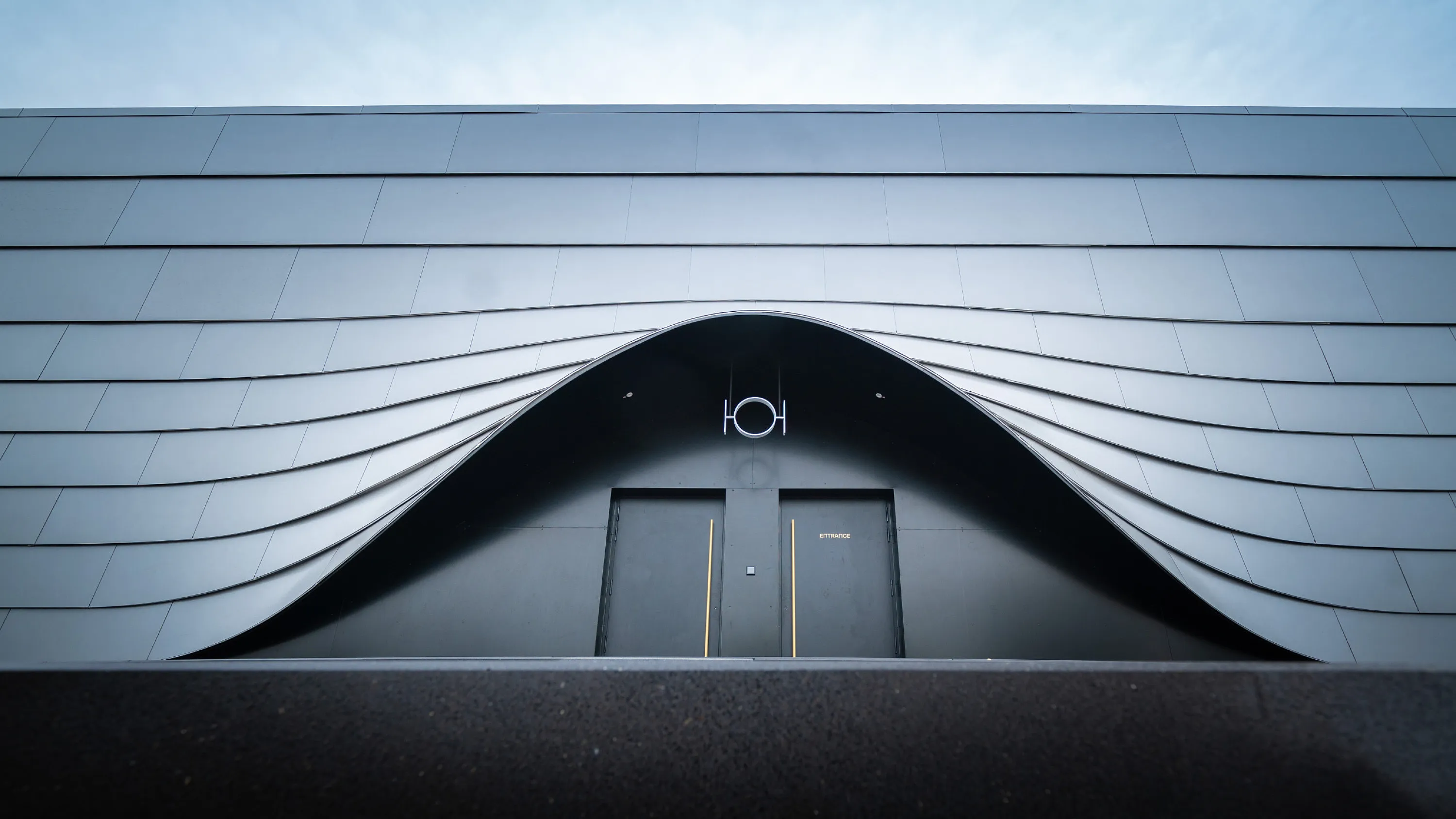 Dark grey, curved metal façade in wave form; in the middle two dark side doors with narrow yellow stripes under an arched entrance, asphalted foreground.