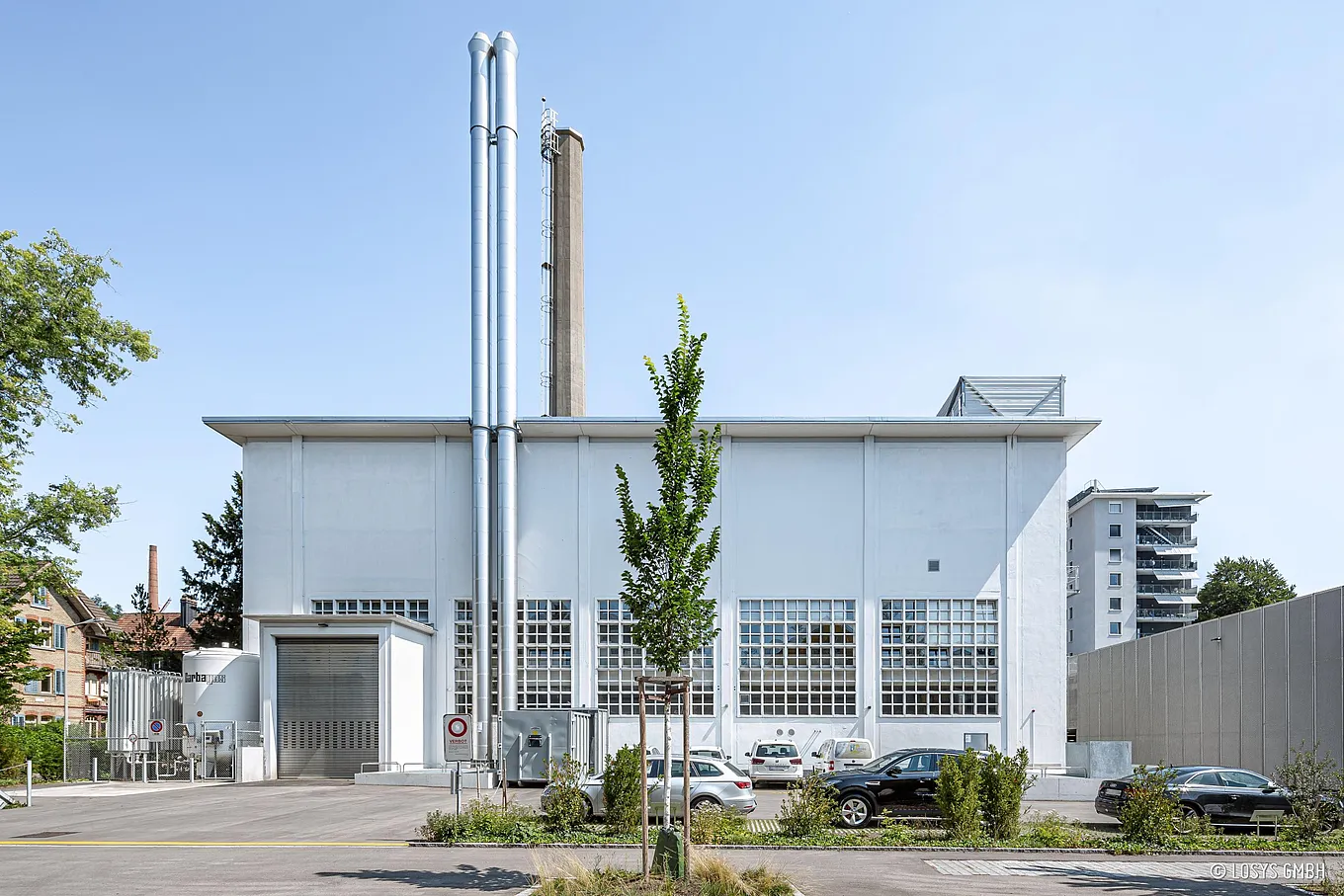 Außenaufnahme einer weißen Industriehalle für eine Notstrom-/Netzersatzanlage: großes Rolltor, vier hohe silberne Abgasrohre, breite Rasterfenster, Parkplätze und Bäume vor dem Gebäude, klarer Himmel.