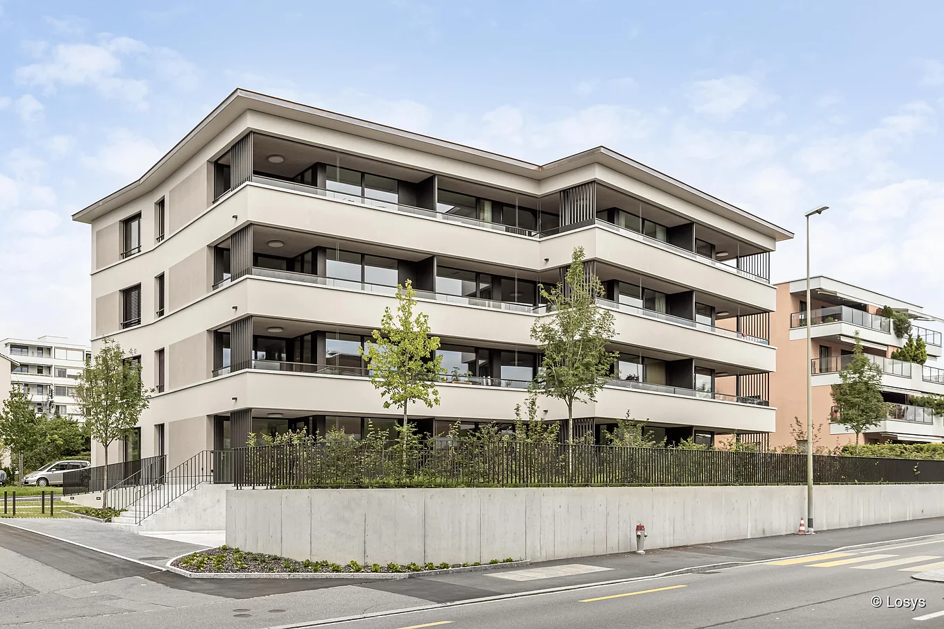Fotografie des viergeschossigen Mehrfamilienhauses «Papillon» in Meilen: hellbeige Fassade mit durchgehenden, zurückgesetzten Balkonen und Glasbrüstungen, dunkle Fensterrahmen, junge Bäume und niedrige Hecken entlang einer erhöhten Betonmauer, Gehweg und Fahrbahn im Vordergrund sowie ein Treppenaufgang zur Hauseingangstür an der linken Gebäudeseite.