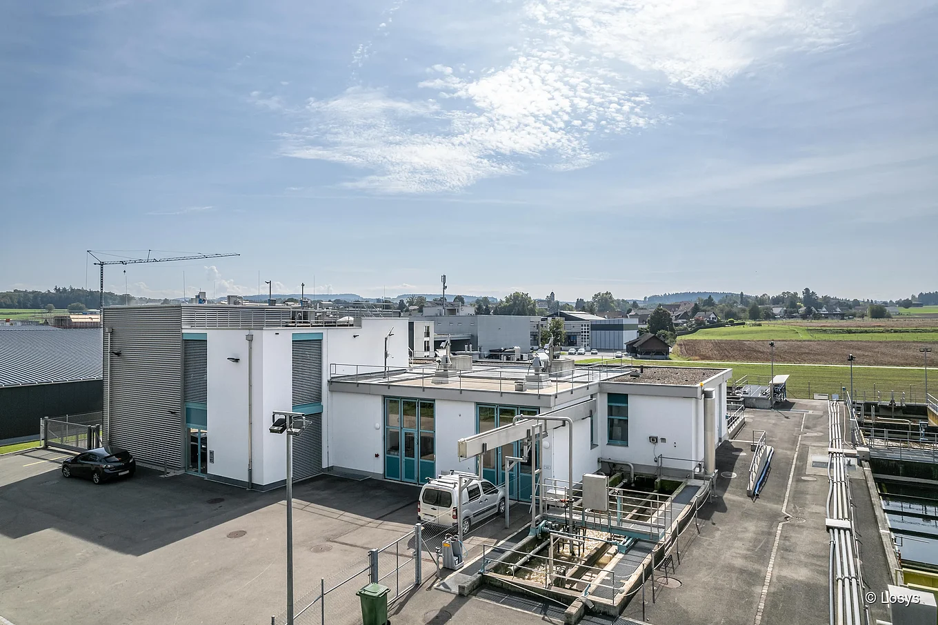 ARA Ellikon: zweigeschossiges Betriebsgebäude einer Abwasserreinigungsanlage mit Flachdächern, Außenklärbecken, Rohrleitungen, Laufstegen, einem weißen Servicewagen und landwirtschaftlicher Umgebung unter klarem Himmel.