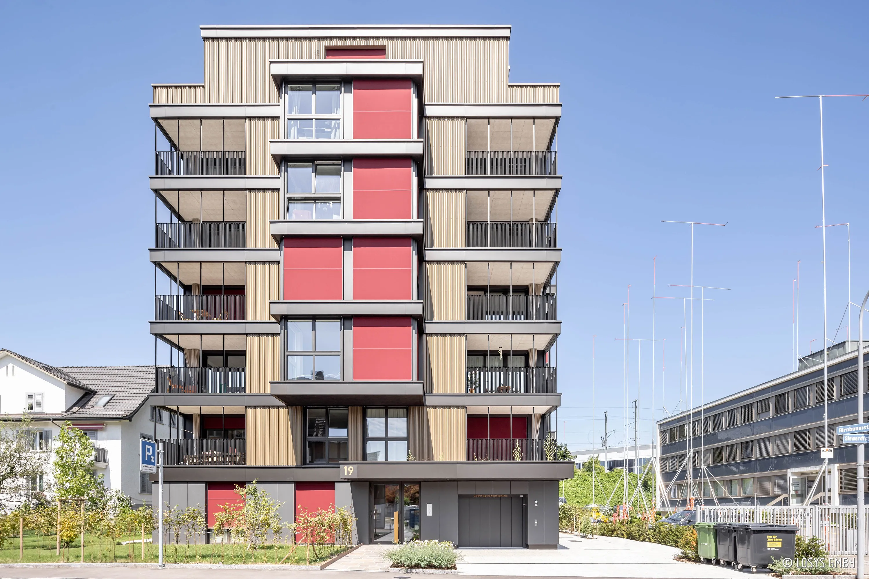 Veduta frontale di un condominio di sei piani con rivestimento verticale della facciata in marrone chiaro e fascia centrale di finestre/pannelli in rosso; balconi su entrambi i lati con ringhiere scure, ingresso principale con numero civico 19 e piccolo giardino anteriore, sullo sfondo un cielo azzurro chiaro.