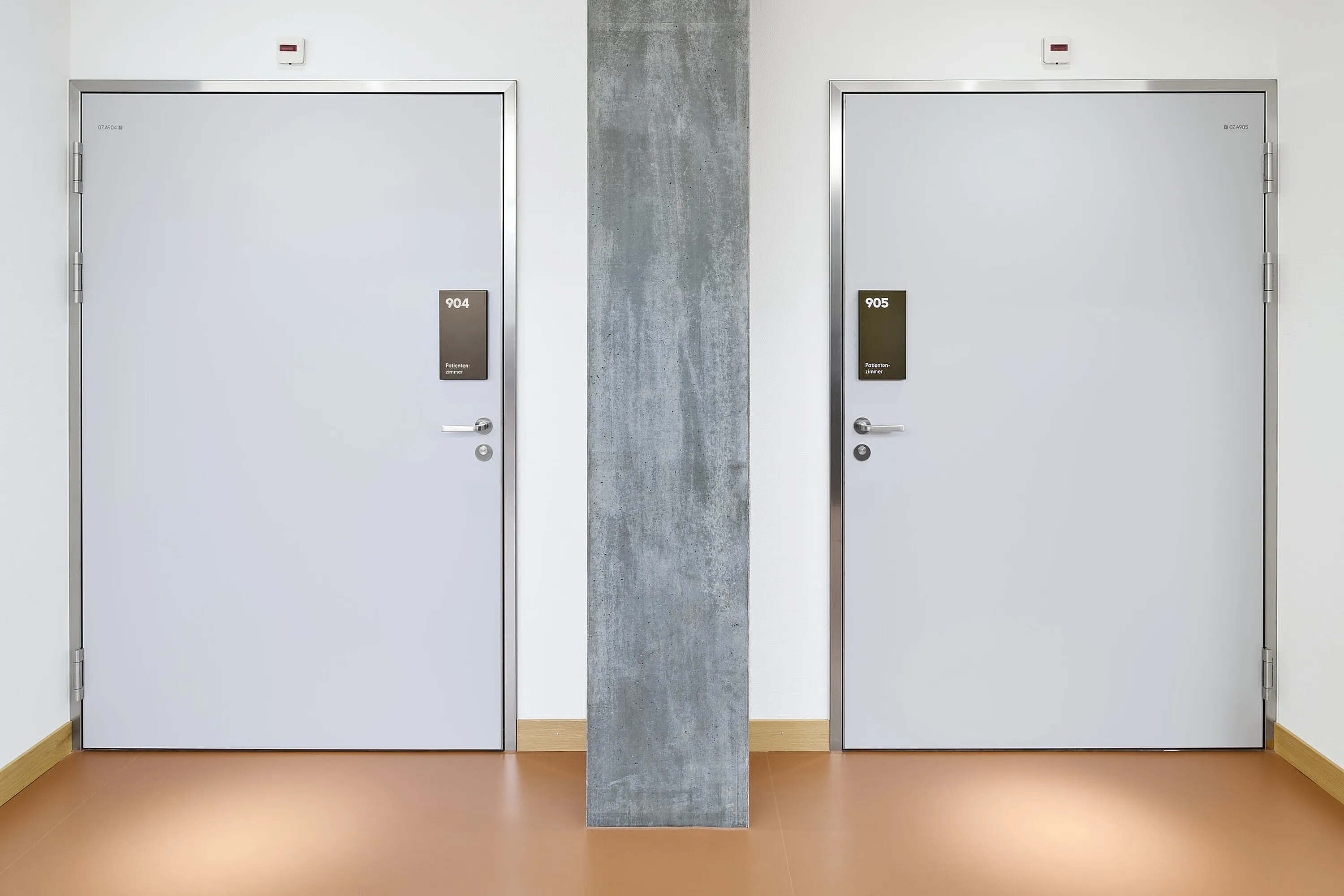 Deux portes intérieures gris clair de même taille avec huisserie en acier argenté et poignées (numéros de pièce 904 et 905) dans un couloir d'hôpital ; au centre de l'image, une colonne en béton, un sol clair et des plinthes en bois.