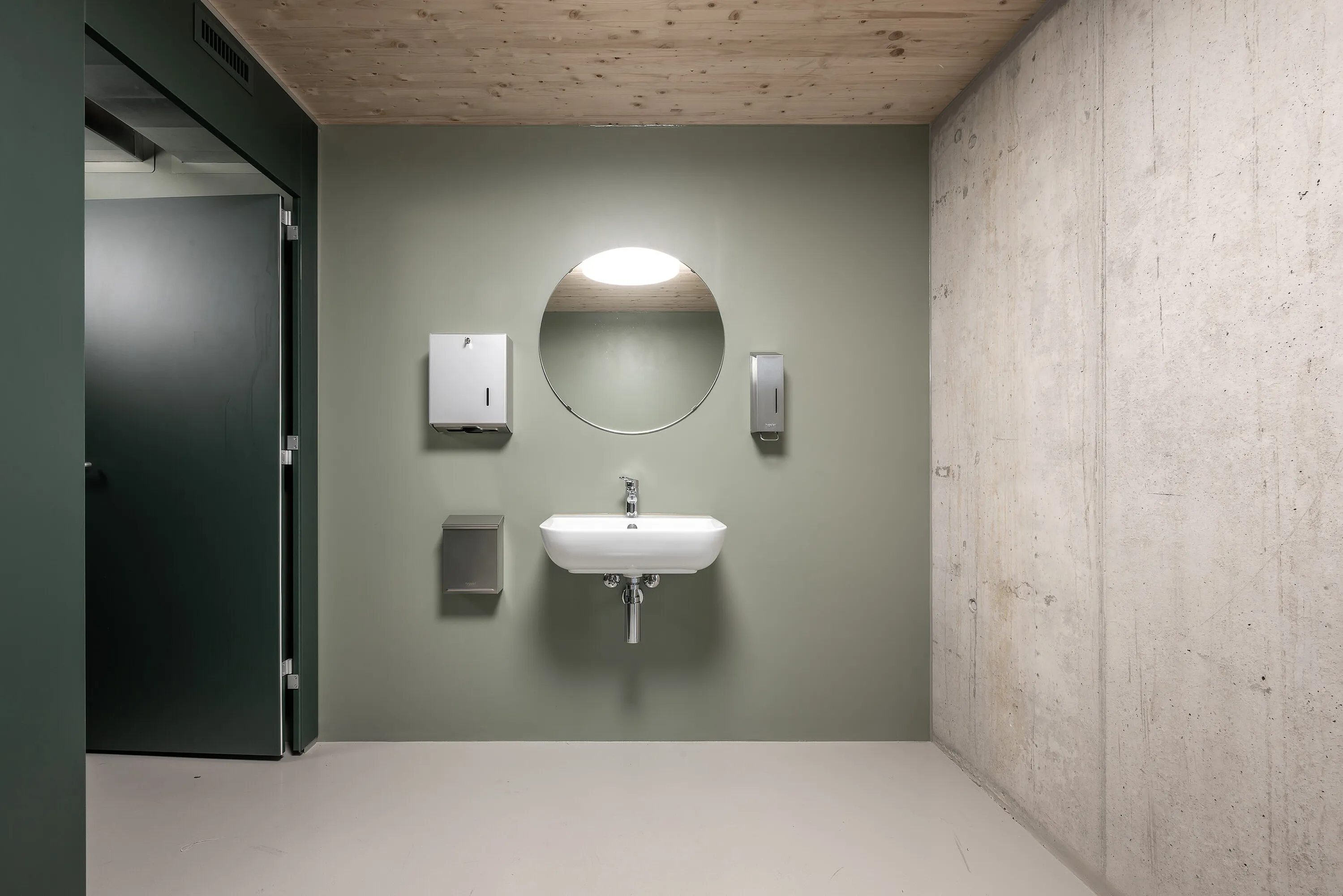 View of a barrier-free sanitary area: matt green wall with round mirror, wall-hung washbasin with visible siphon, soap and paper towel dispenser and waste bin; dark interior door open on the left, raw concrete wall, light-coloured floor and wooden ceiling on the right.