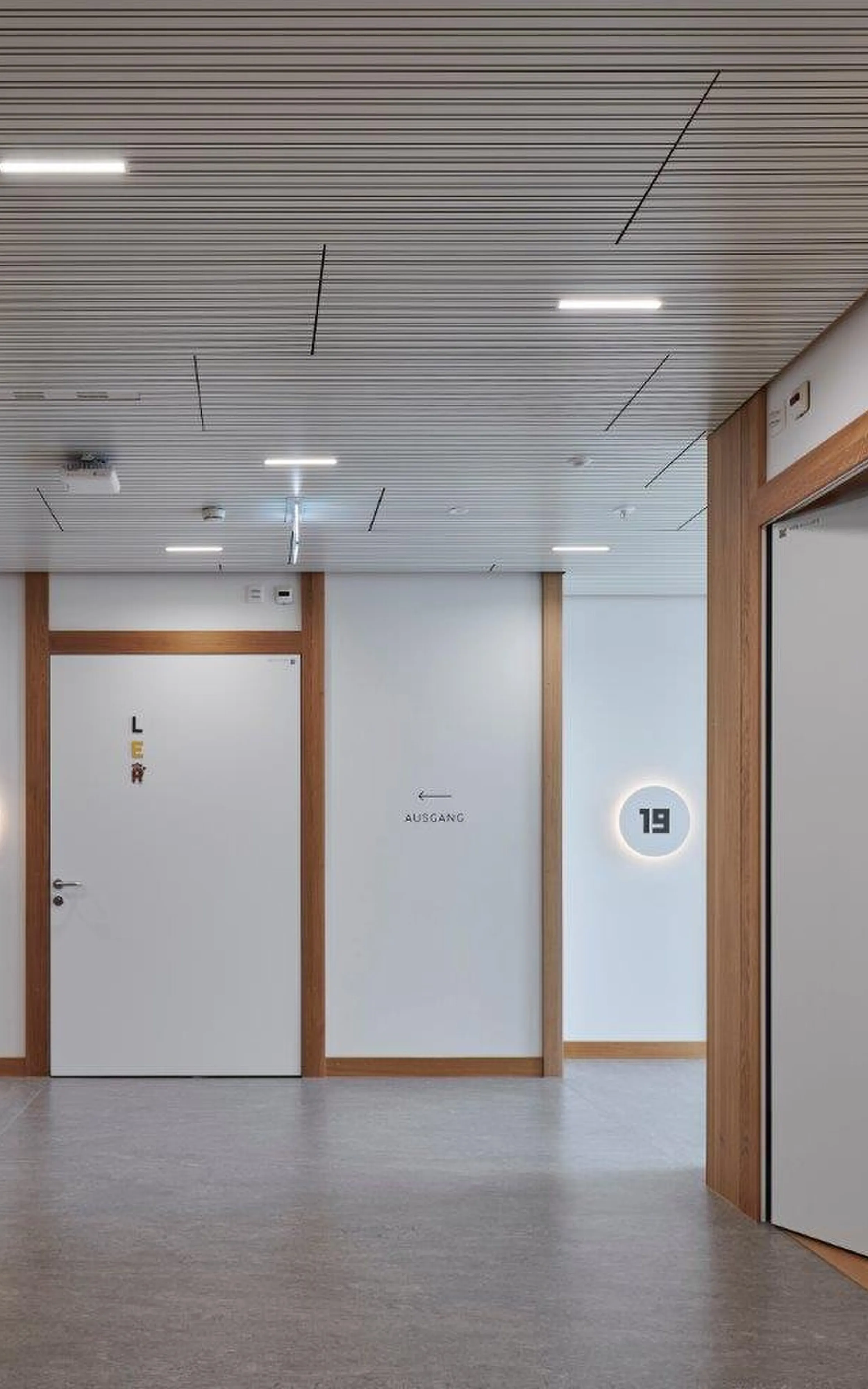 Couloir de clinique clair avec un sol gris clair, des cadres en bois sur les murs et le plafond, plusieurs portes intérieures blanches avec des huisseries en bois, des numéros de chambre ronds éclairés (18-20) et une flèche 'sortie' sur le mur.