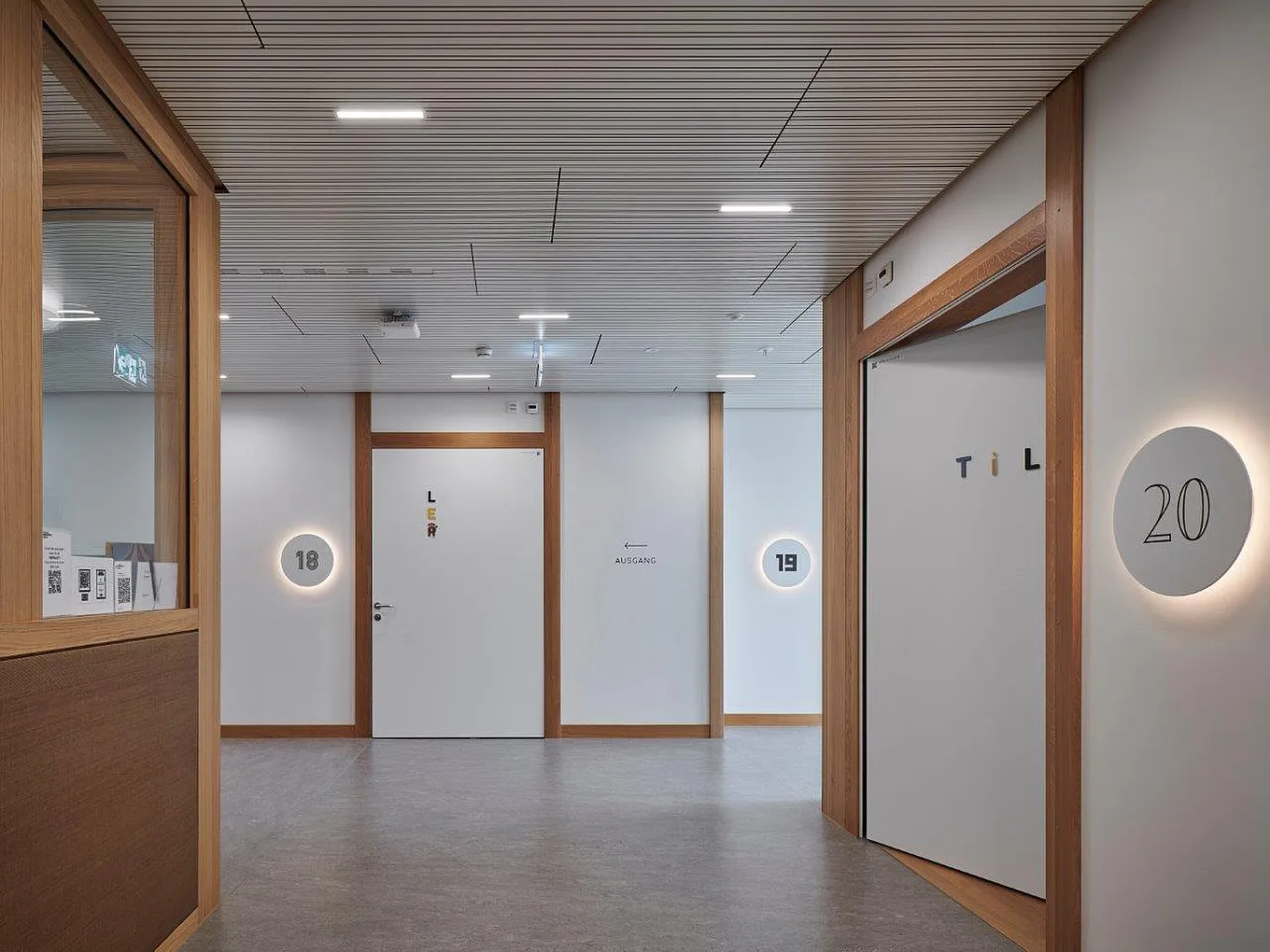 Couloir de clinique clair avec un sol gris clair, des cadres en bois sur les murs et le plafond, plusieurs portes intérieures blanches avec des huisseries en bois, des numéros de chambre ronds éclairés (18-20) et une flèche 'sortie' sur le mur.