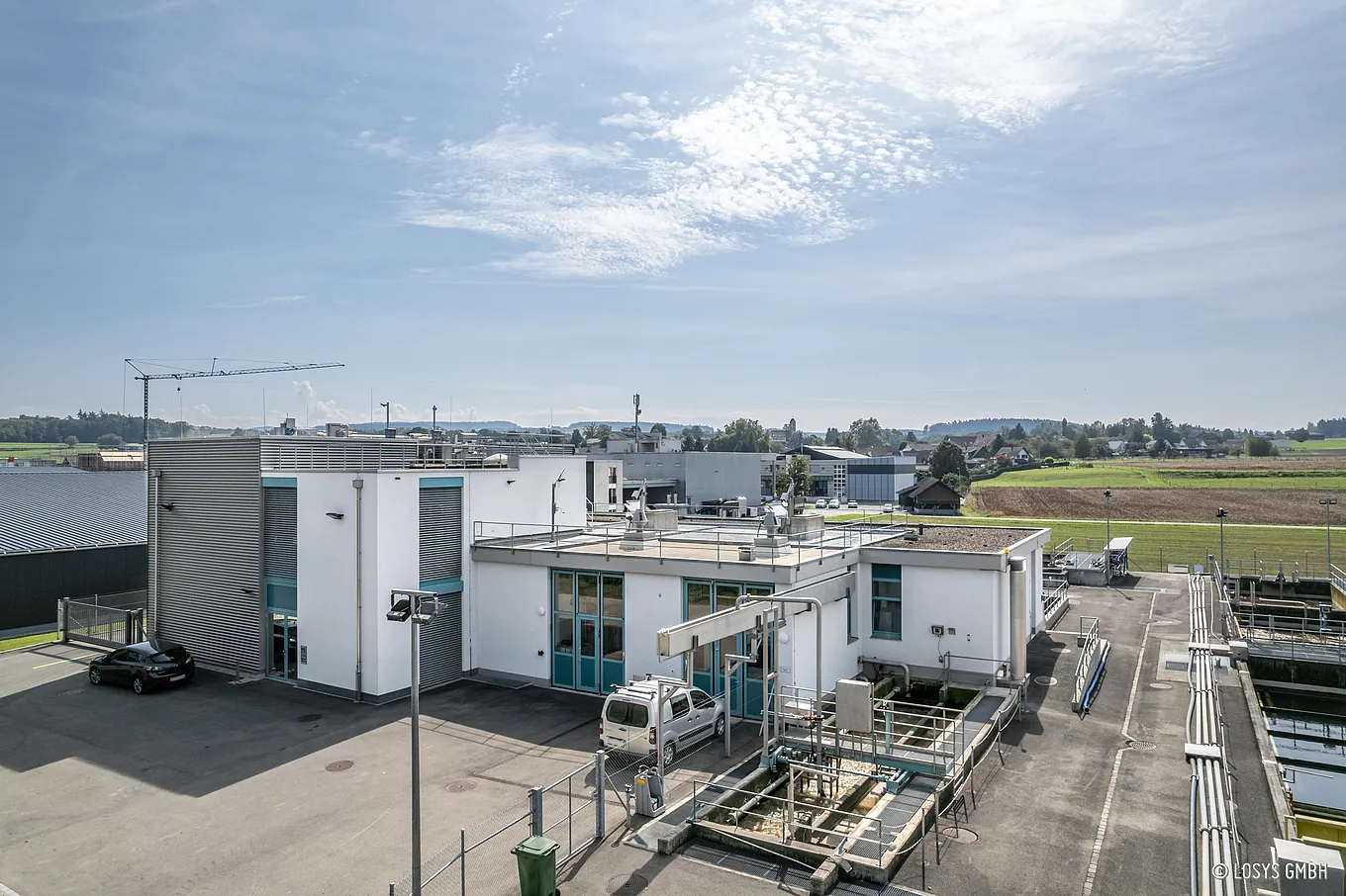 Vue aérienne de la STEP d'Ellikon : un bâtiment d'exploitation blanc avec des portes vitrées/doubles portes turquoises, des bassins d'épuration adjacents, des canalisations, des passerelles et des balustrades, une voiture de service dans la cour et des champs en arrière-plan.