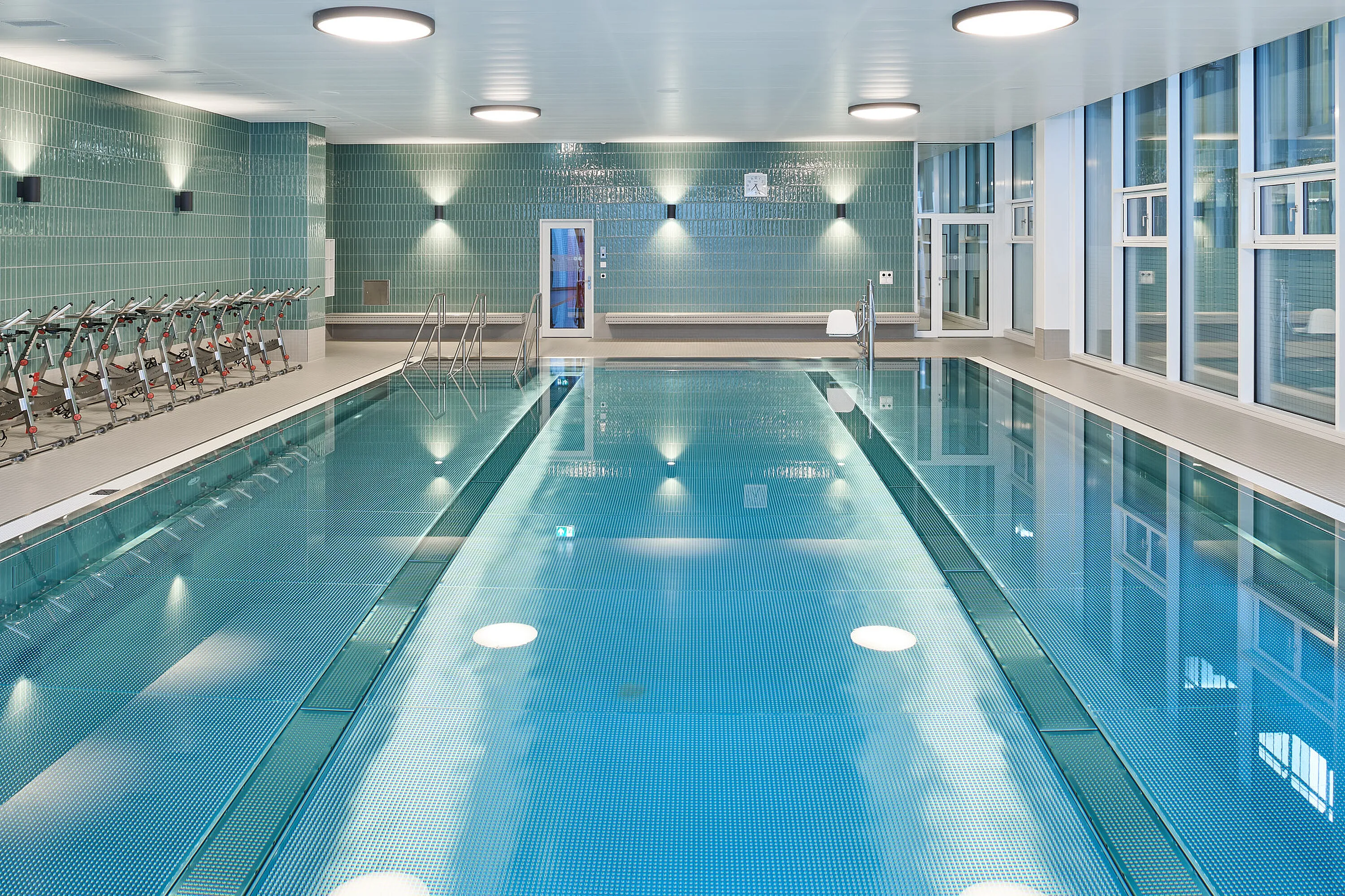 Therapiebad innen: rechteckiges Therapiebecken mit Treppe und Handläufen, Pool‑Lift, Reihe von Unterwasser‑Therapierädern an der Seite, grünliche Wandfliesen und große Fensterfront.