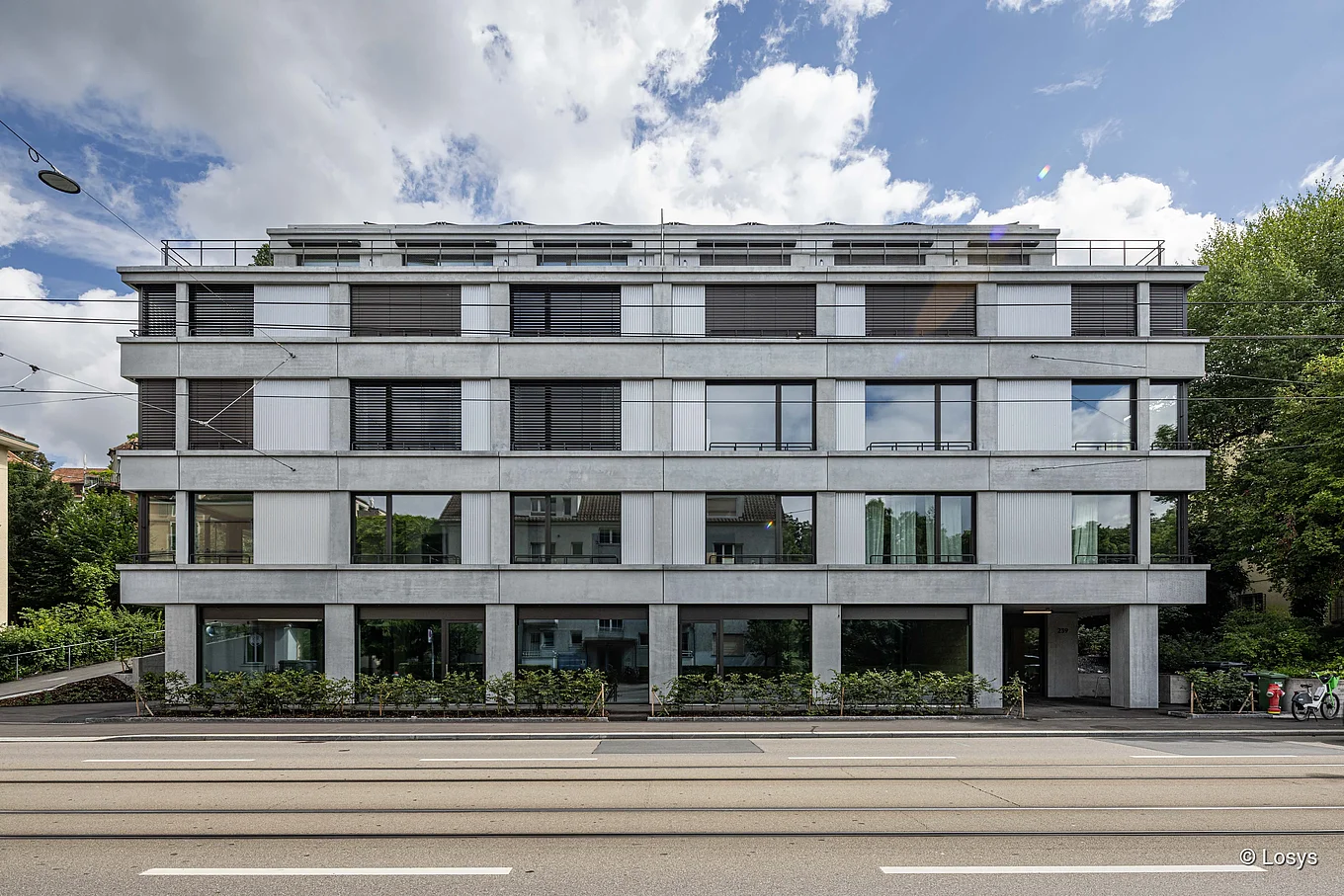 Vorderansicht der Wohnüberbauung Forchstrasse in Zürich: kubischer, viergeschossiger Betonbau mit horizontalen Fensterbändern und grossflächiger Verglasung, teils mit geschlossenen Lamellenjalousien. Erdgeschoss mit Glasfronten und seitlichem Hauseingang rechts, zurückgesetztes Dachgeschoss mit Terrasse. Strassenbahngleise und Gehweg im Vordergrund, Bäume an den Gebäudeenden.