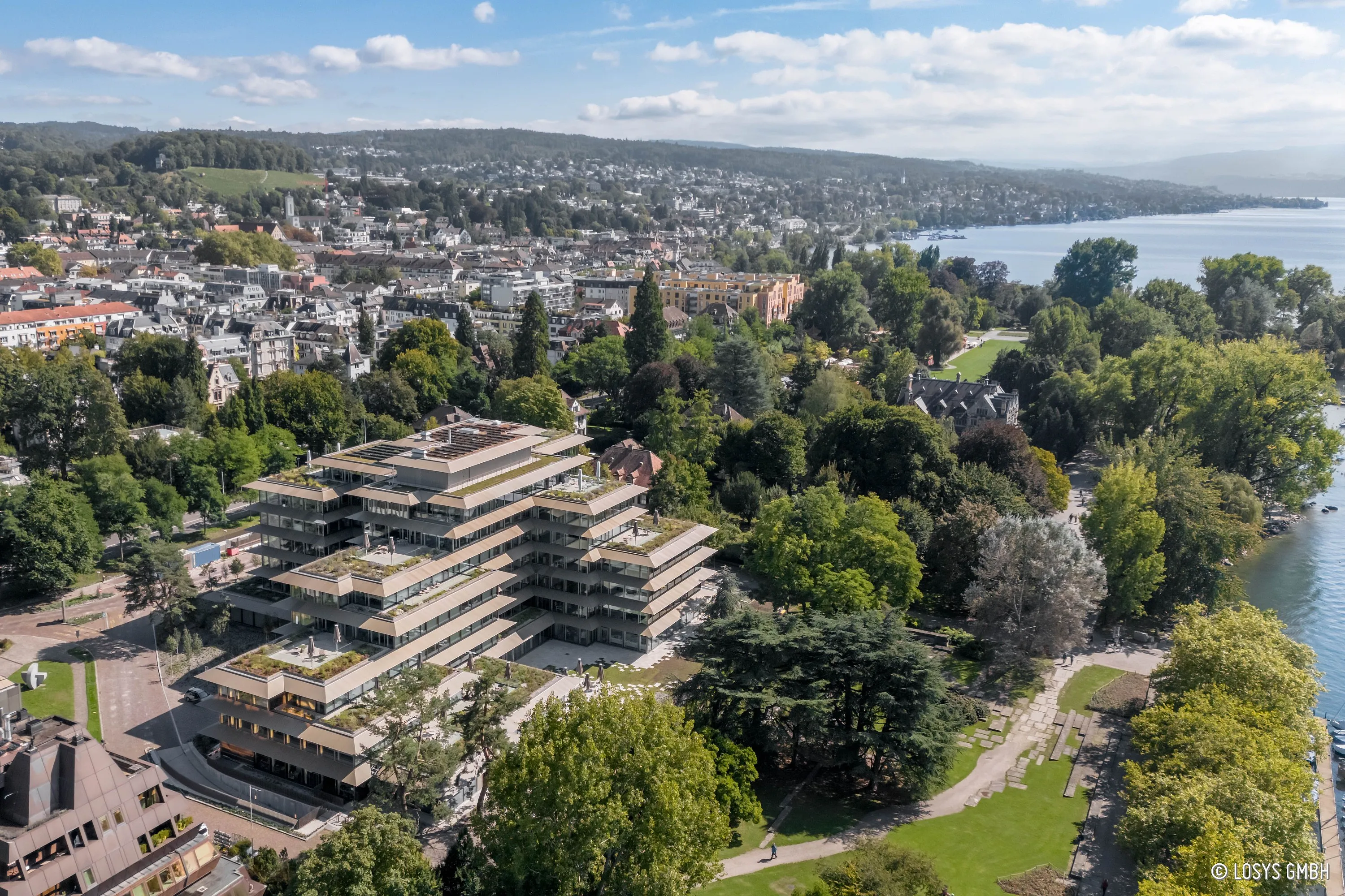 Bird's-eye view of a terraced commercial building on Bellerivestrasse in Zurich with stepped, green roof terraces, adjoining park and lakeside promenade.