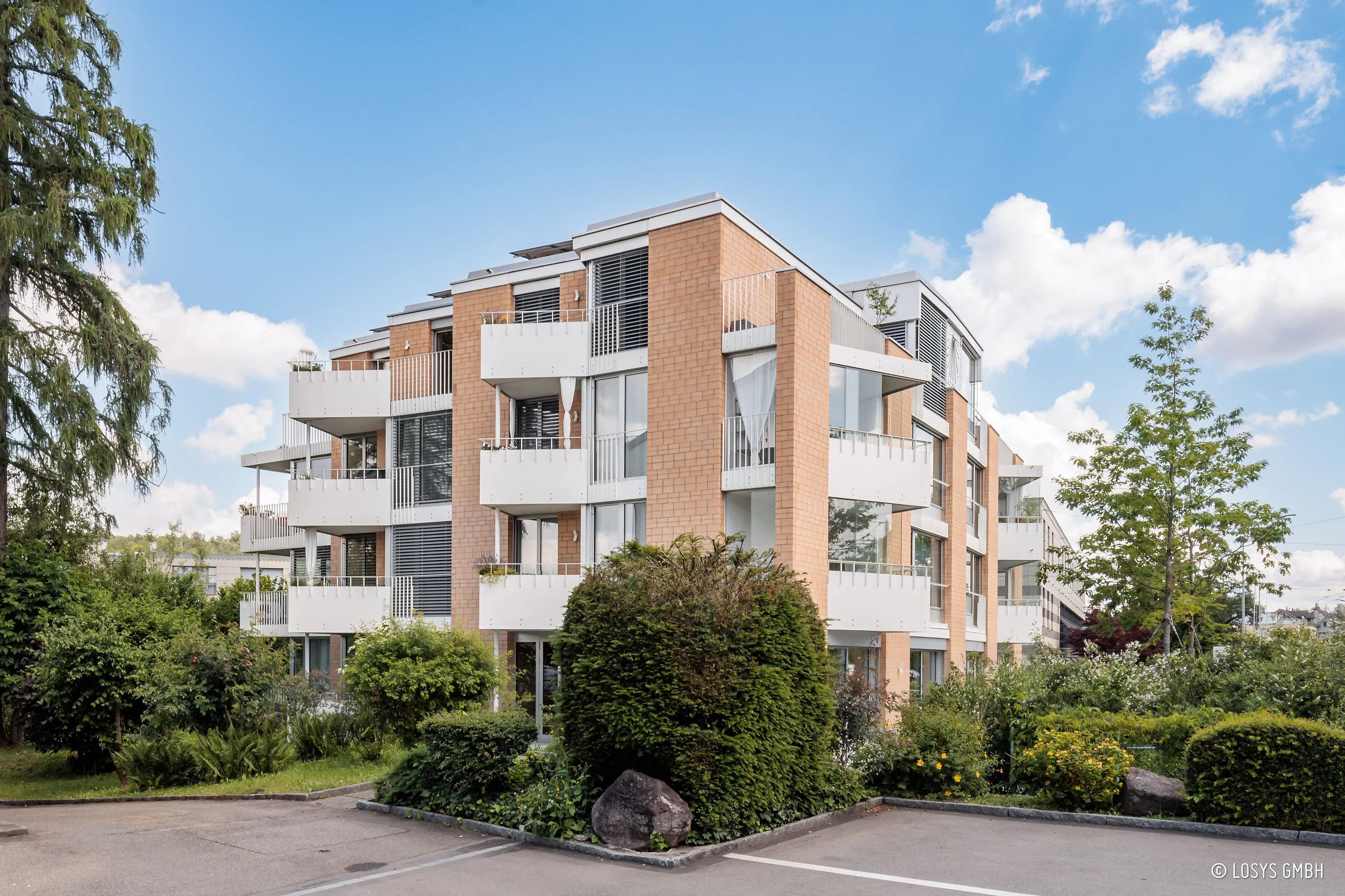 Vorderansicht eines viergeschossigen Mehrfamilienhauses mit hellbrauner Klinkerfassade, gestaffelten weißen Balkonen und großflächigen Glasfenstern/-türen; Vorgarten mit Sträuchern, Parkfläche im Vordergrund und blauem Himmel.