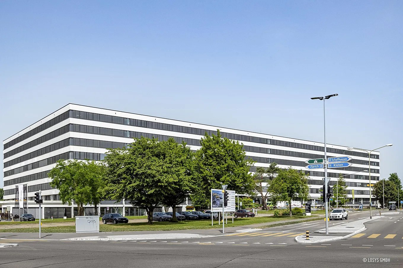 Referenzbild: Aufstockung des Geschäftshauses Balsberg. Breites, längliches mehrgeschossiges Bürogebäude mit abwechselnden weißen und dunklen horizontalen Fensterbändern; im Vordergrund Bäume, parkende Autos, Fußgängerüberwege, Verkehrsampeln und Wegweiser an einer Straßenkreuzung, klarer blauer Himmel.