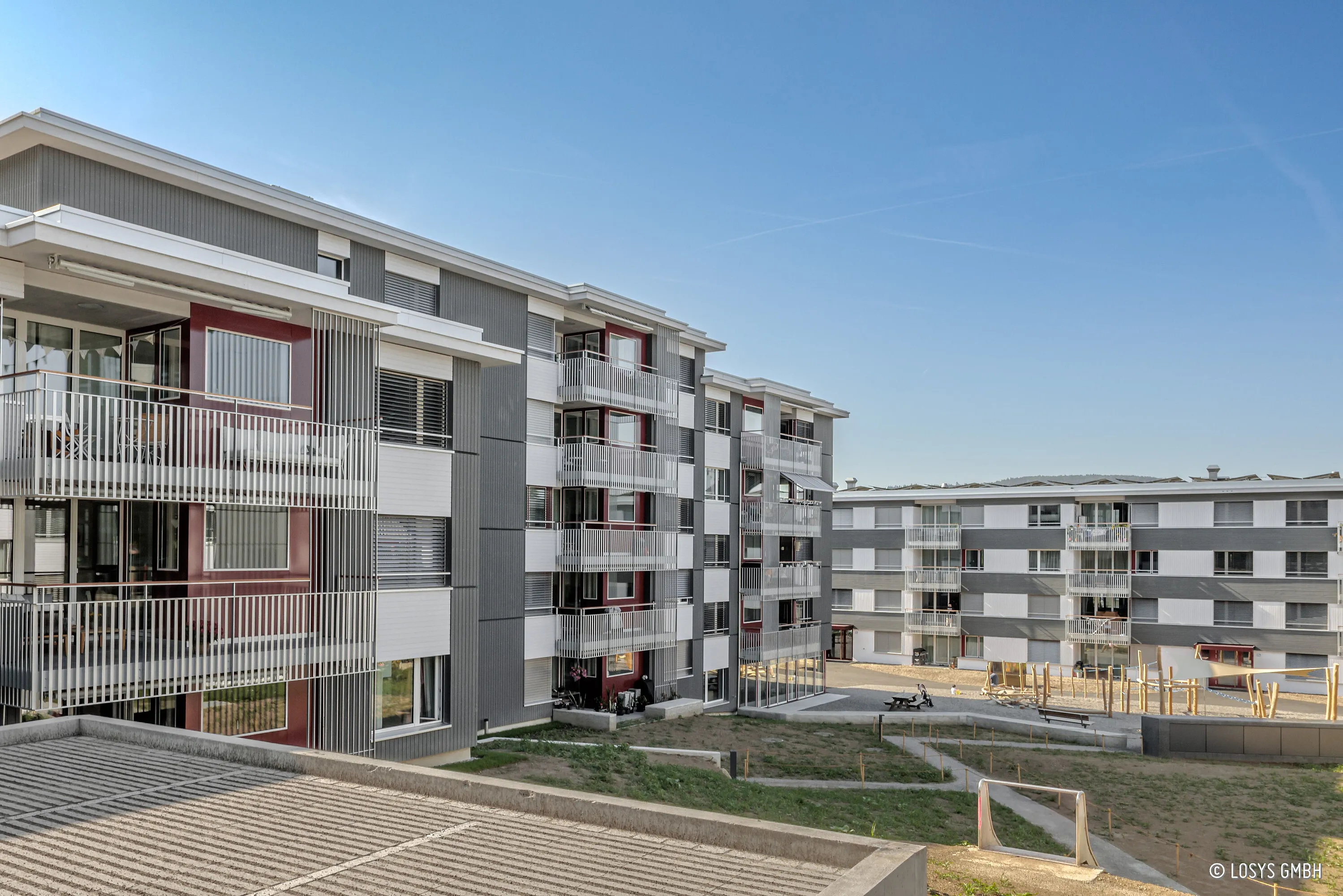 Plusieurs immeubles d'habitation modernes de quatre étages avec des balustrades de balcon blanches, des accents de façade gris et rouge ; au premier plan, niveau de parking, cour intérieure avec des chemins et une petite aire de jeux sous un ciel bleu.