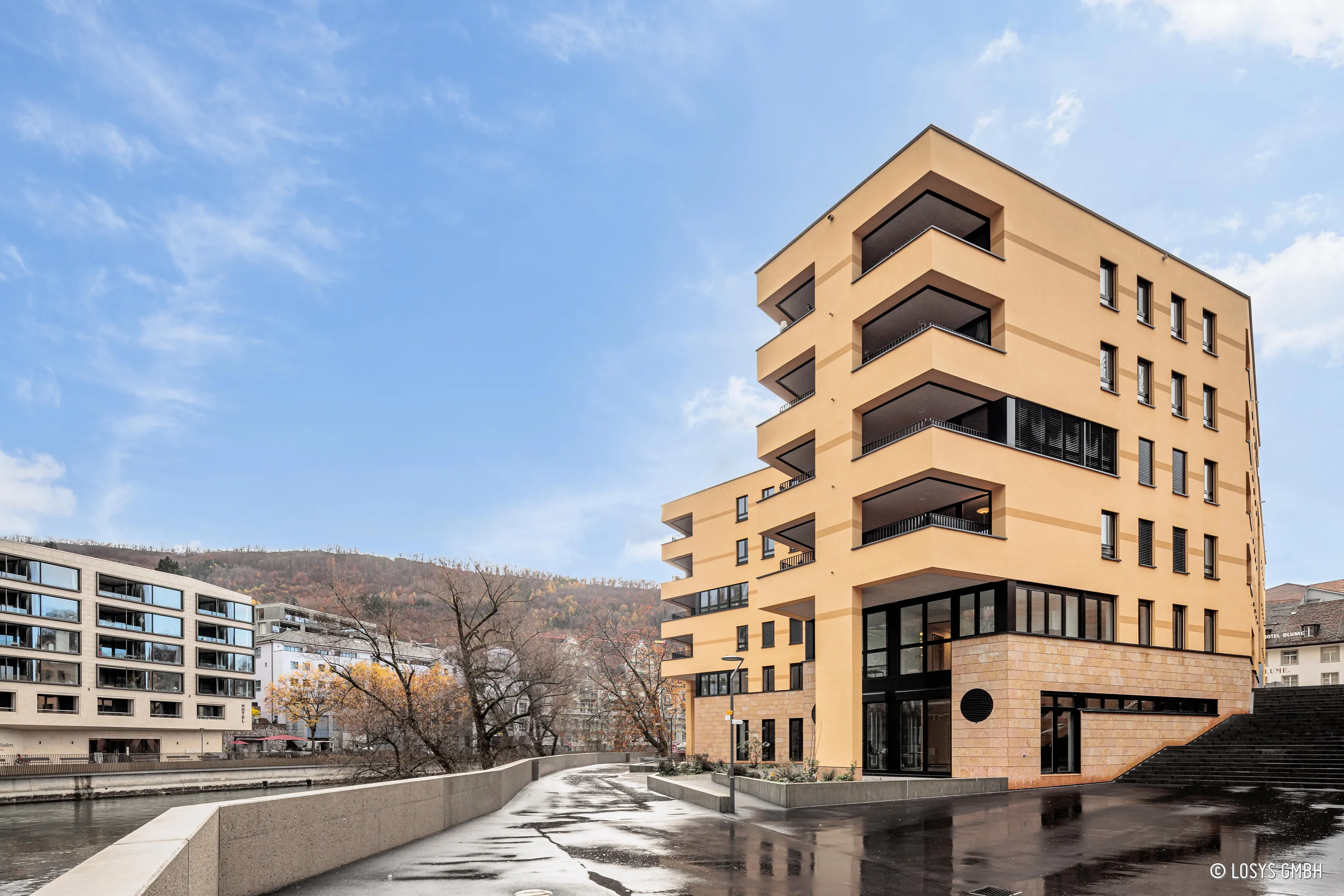 Photo extérieure du centre médical jaune et beige "Fortyseven" au bord de la rivière : bâtiment moderne à plusieurs étages avec balcons en retrait, grandes baies vitrées, hall d'entrée, escaliers et promenade mouillée par temps clair.