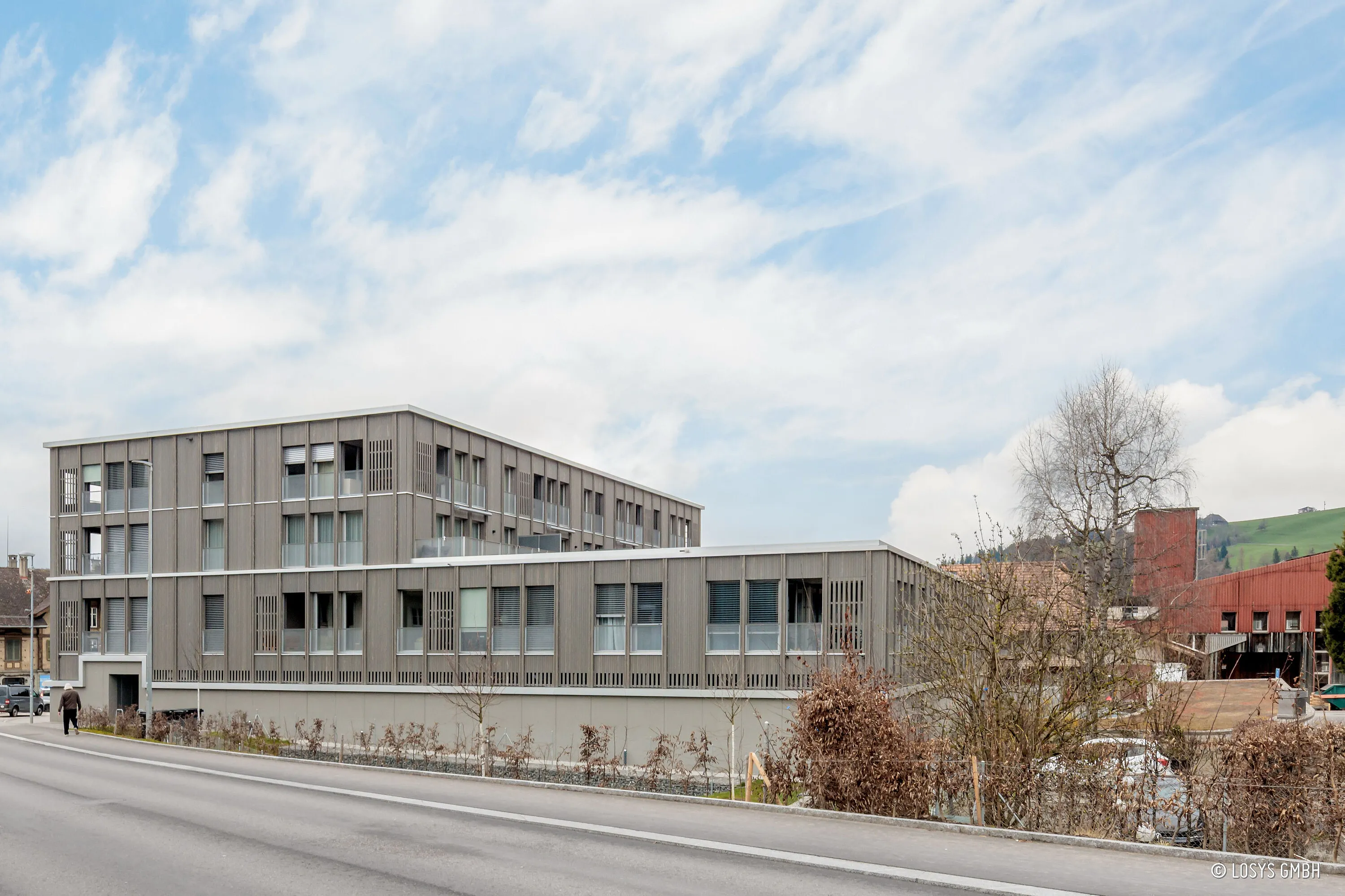Exterior view of the new building at Ilfiskreisel in Langnau: modern three-storey building with a light grey, vertically structured façade, regular windows and loggias; road, pavement and individual trees in the foreground.