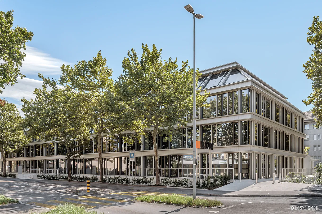 Dreigeschossiges, modernes Bürogebäude mit durchgehender Glasfassade und sichtbarem BKW-Logo; vor dem Gebäude Bäume, Gehweg, Laterne und barrierefreier Eingangsbereich mit Rampe.