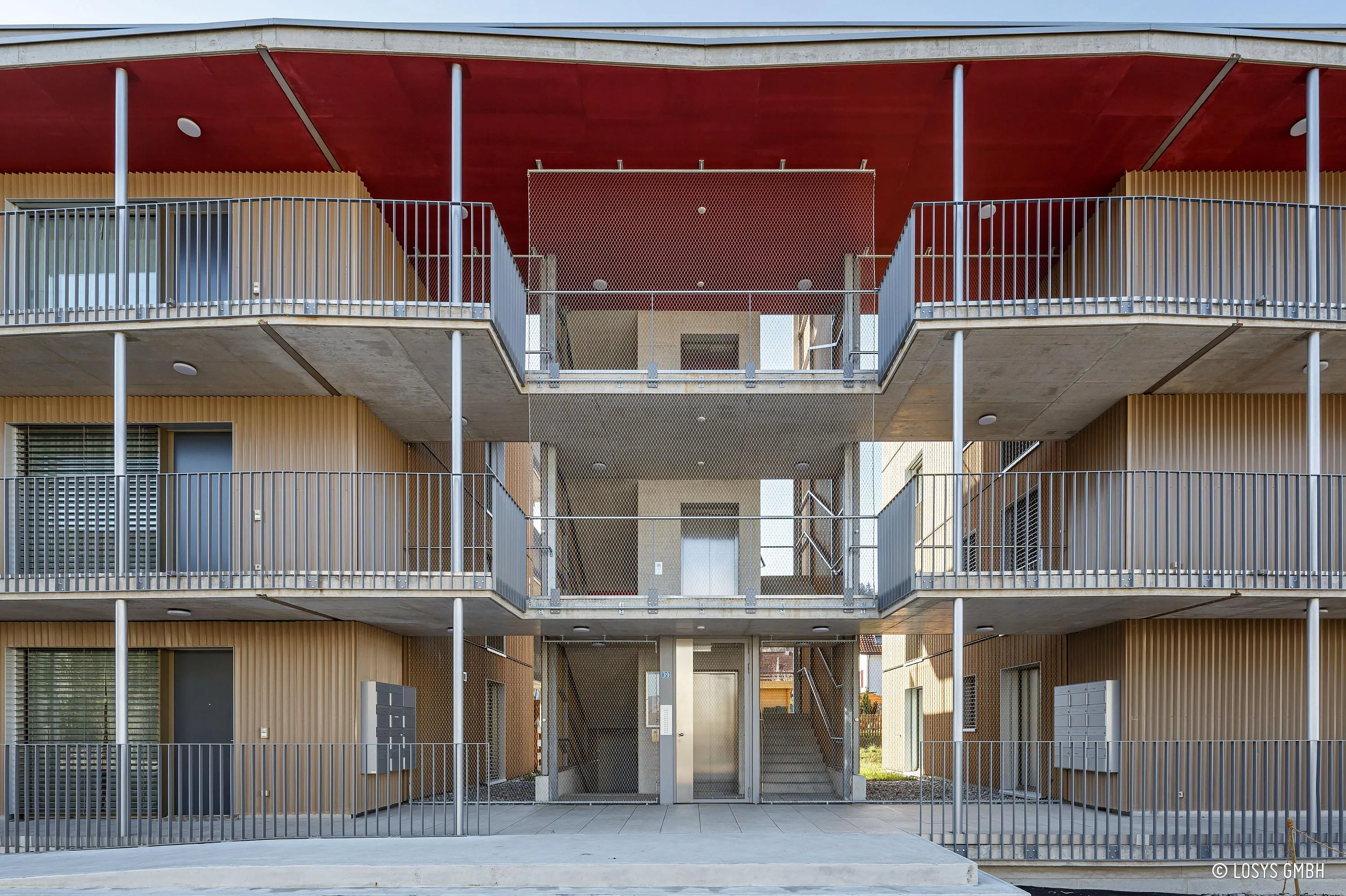 Dreigeschossige Wohnfassade mit zentralem Treppenhaus, Glastür, Betonbalkonen und heller Holzverkleidung.